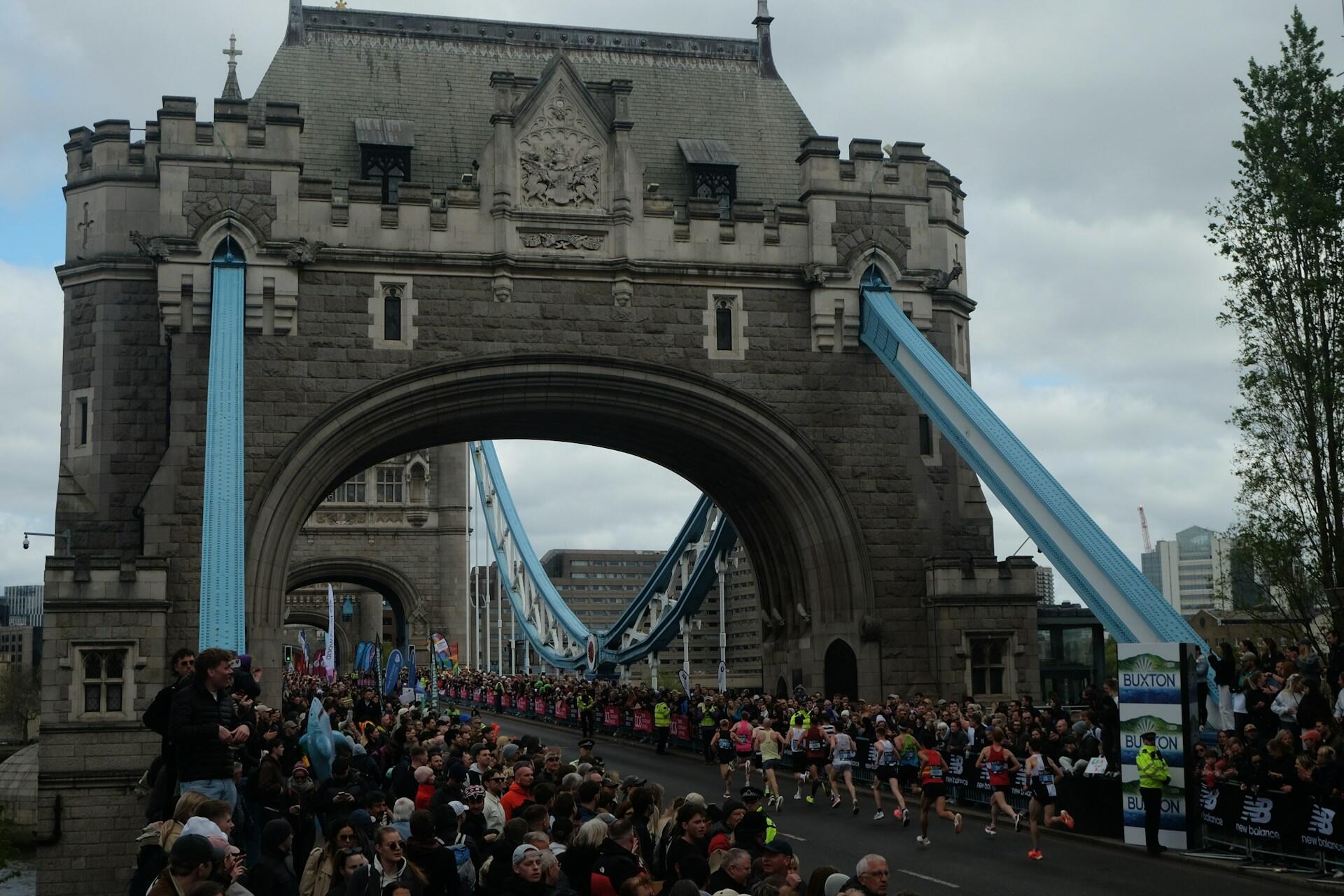 Runners crossing Tower Bridge during a marathon race with crowds of spectators watching.