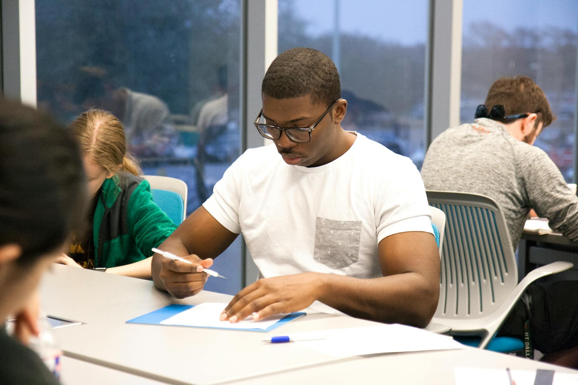 Students focused on writing and studying at a classroom table with large windows and blue chairs.