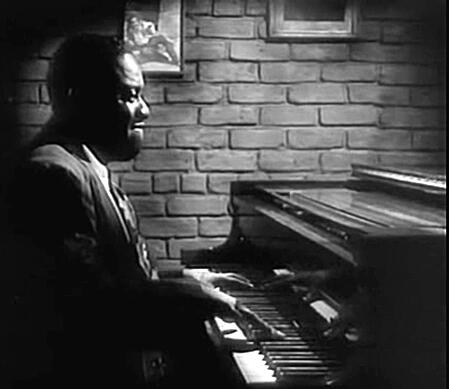 Black and white profile view of a man in a suit intently playing an upright piano in front of a brick wall.