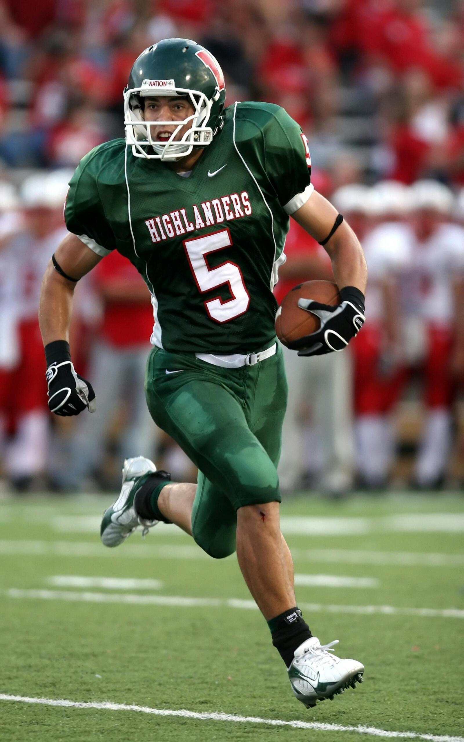 A football player in a green Highlanders jersey with number 5 sprints on a field, holding a ball, amid a blurred crowd in the background.