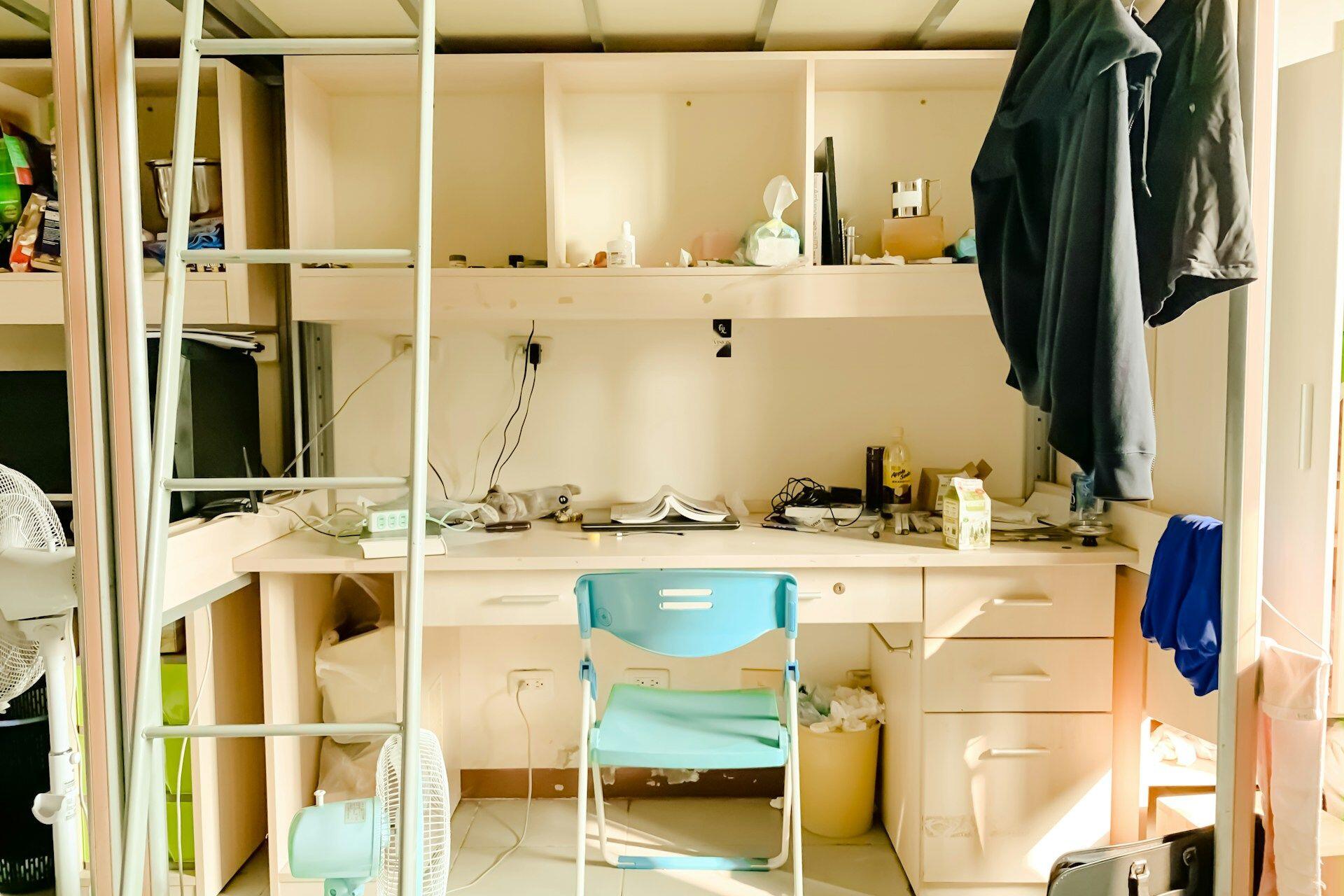 Study desk under a loft bed with a blue chair, cluttered with books, devices, clothes hanging, and various items on shelves and desk.