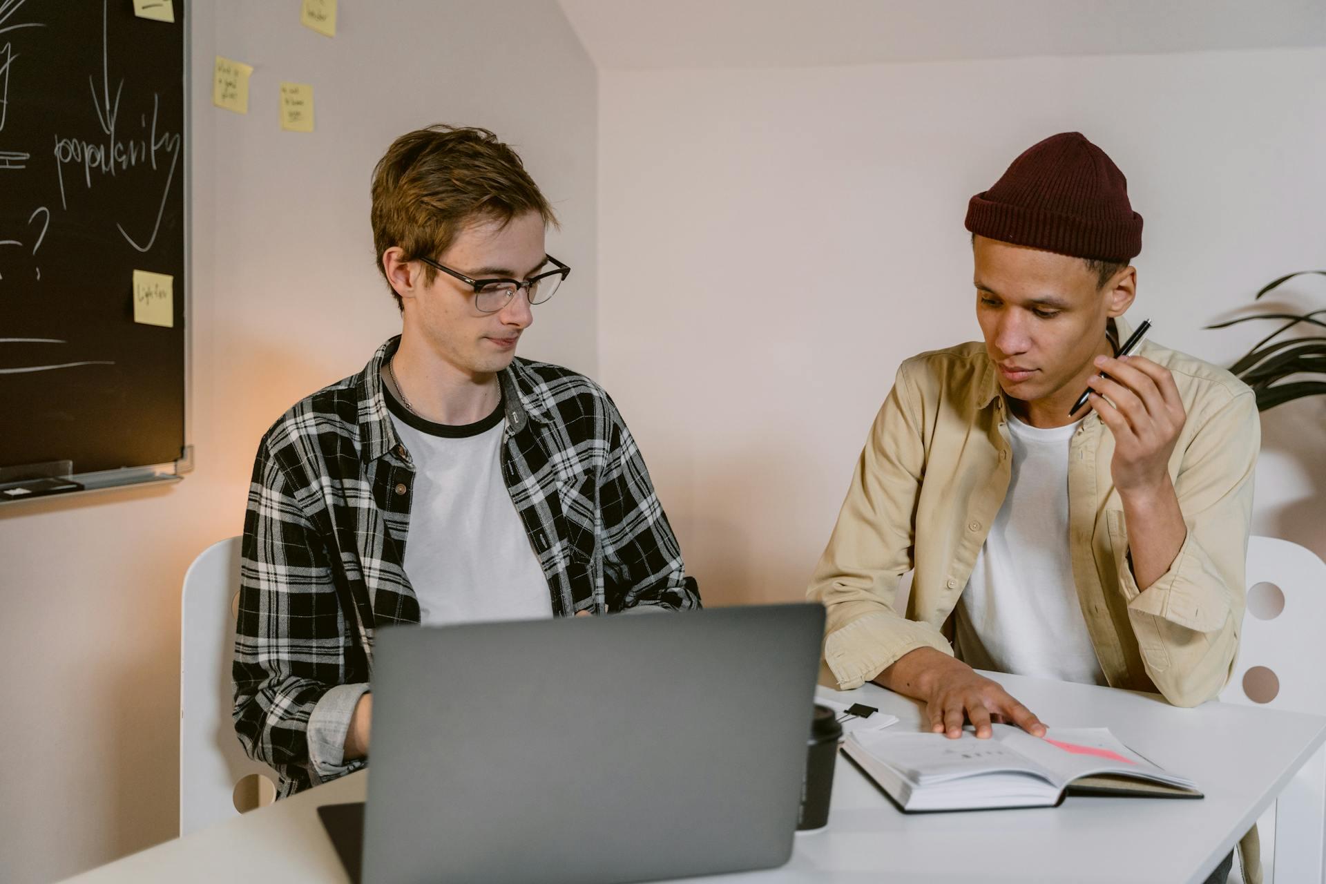 Two young men discussing notes with laptop and open book in a study room with sticky notes on the wall and a blackboard.
