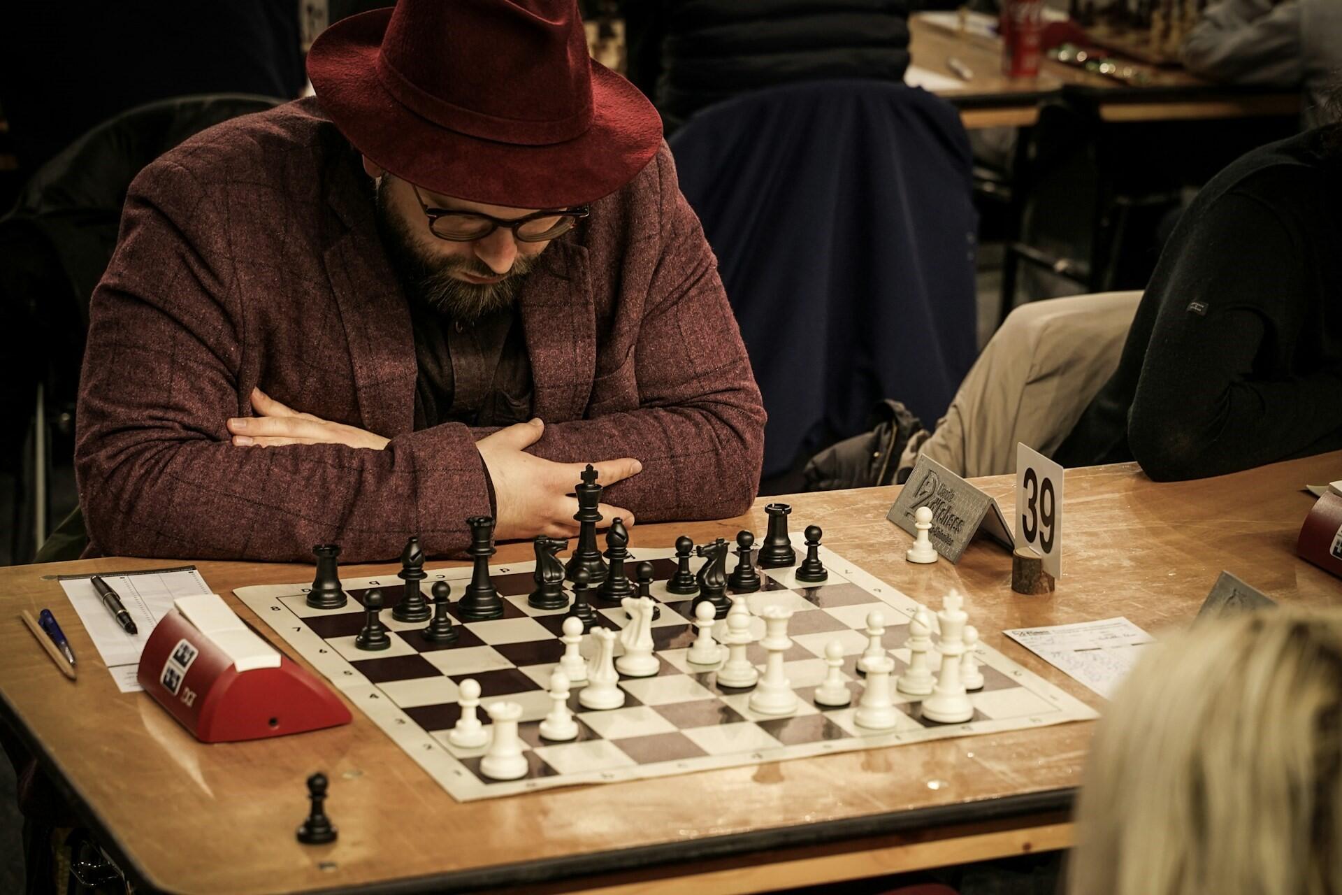 A bearded chess player wearing a red hat studies a chess position during a tournament game while seated at a board with pieces mid-game.