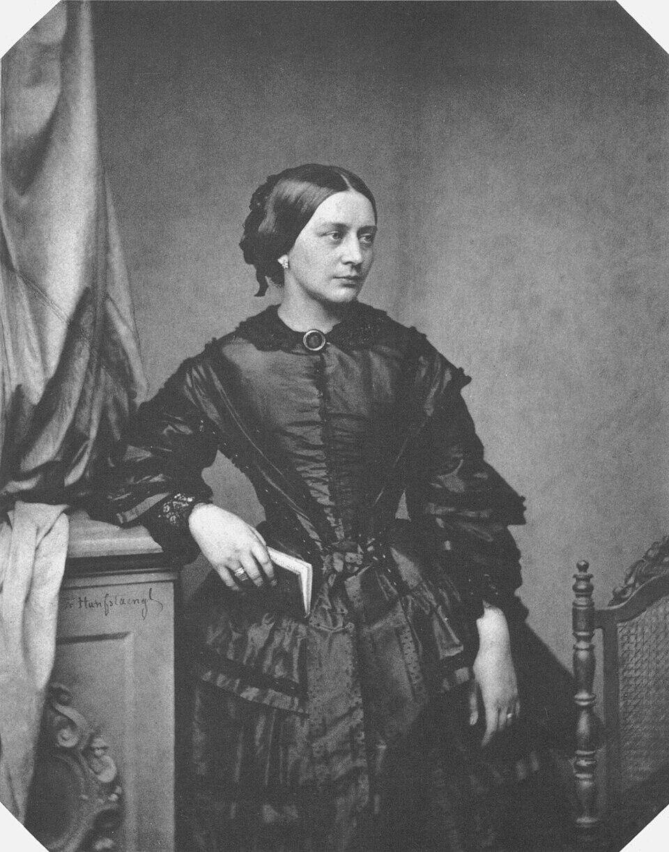 Monochrome portrait of a young woman in a dark, voluminous dress standing beside a draped pedestal while holding a small book.