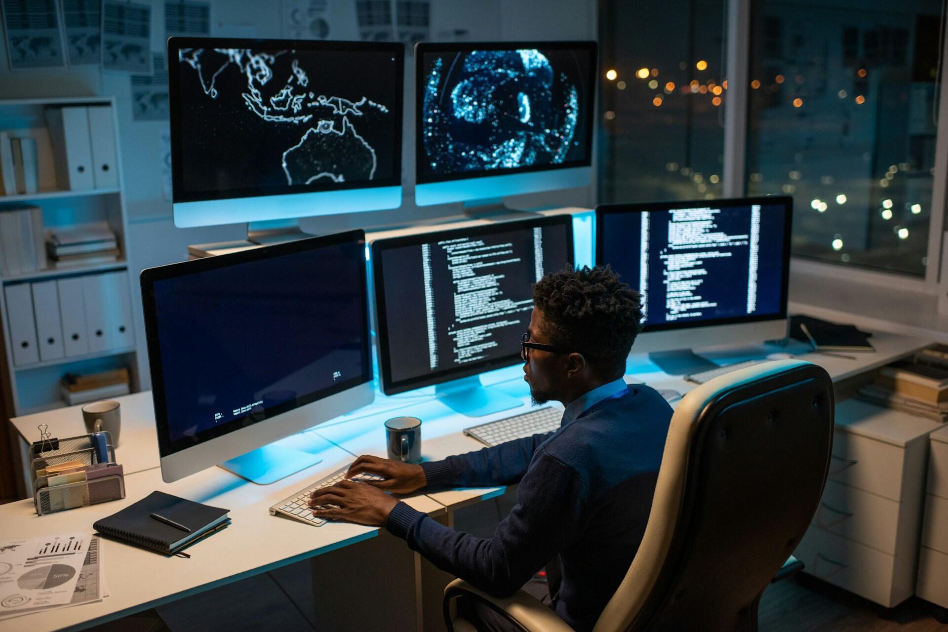 A software engineer working at a desk with six computer monitors displaying code in an office at night.
