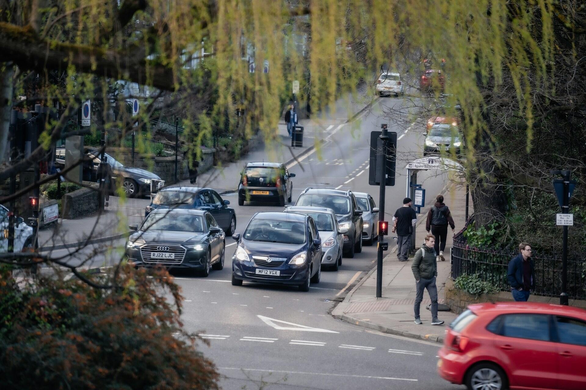Busy city street with cars waiting at a red light and pedestrians walking on the sidewalk near trees and greenery.