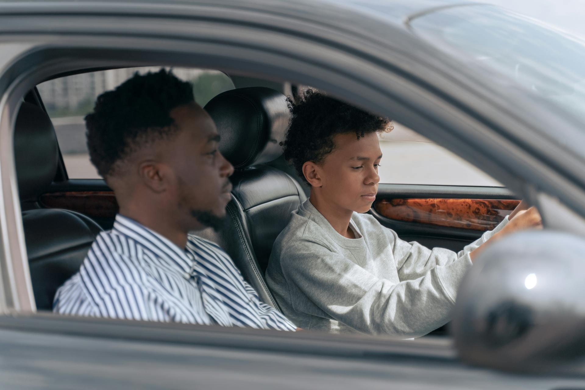 Two young men sitting inside a car, one driving and the other in the passenger seat, visible through the open window.