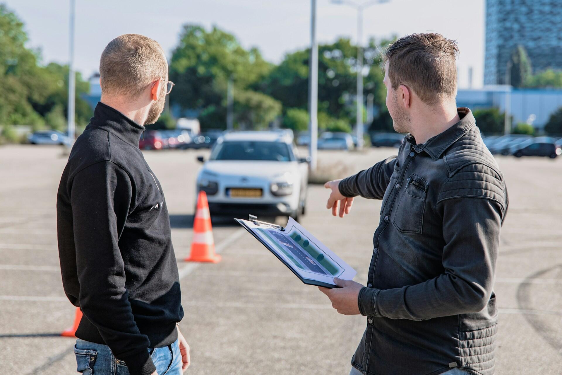 Two men discuss a driving course on a parking lot, with one pointing towards a white car near traffic cones.