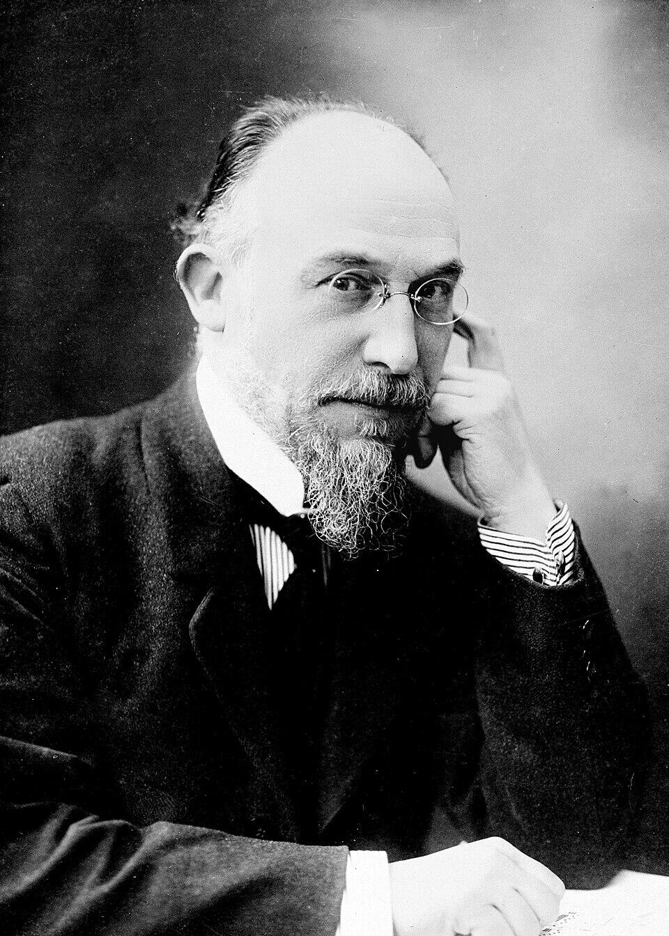 Black and white portrait of a balding man with a prominent beard and round glasses, posed thoughtfully with his hand near his face.