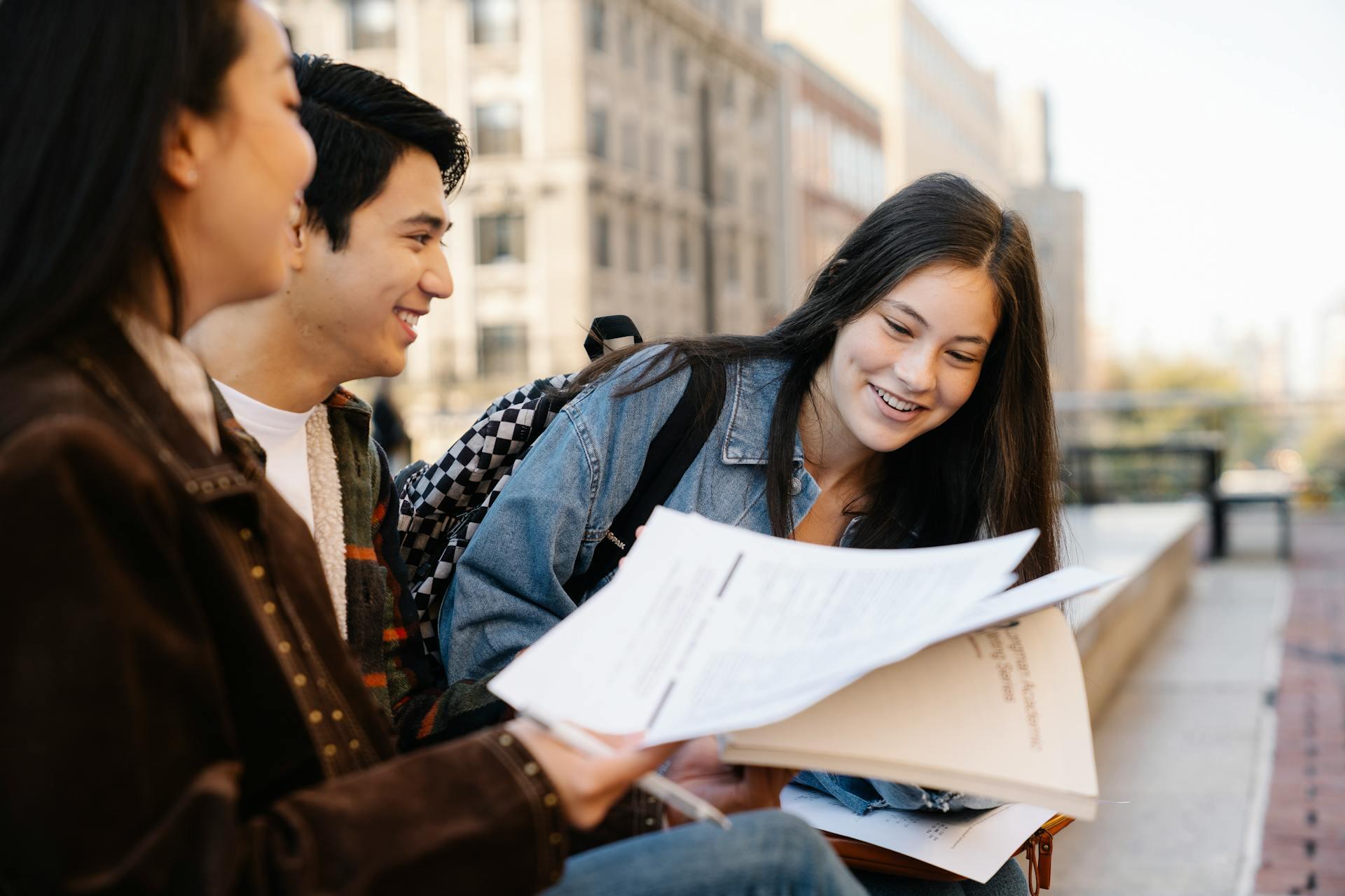 Three students outdoors reviewing and discussing documents, with a cityscape background on a sunny day.