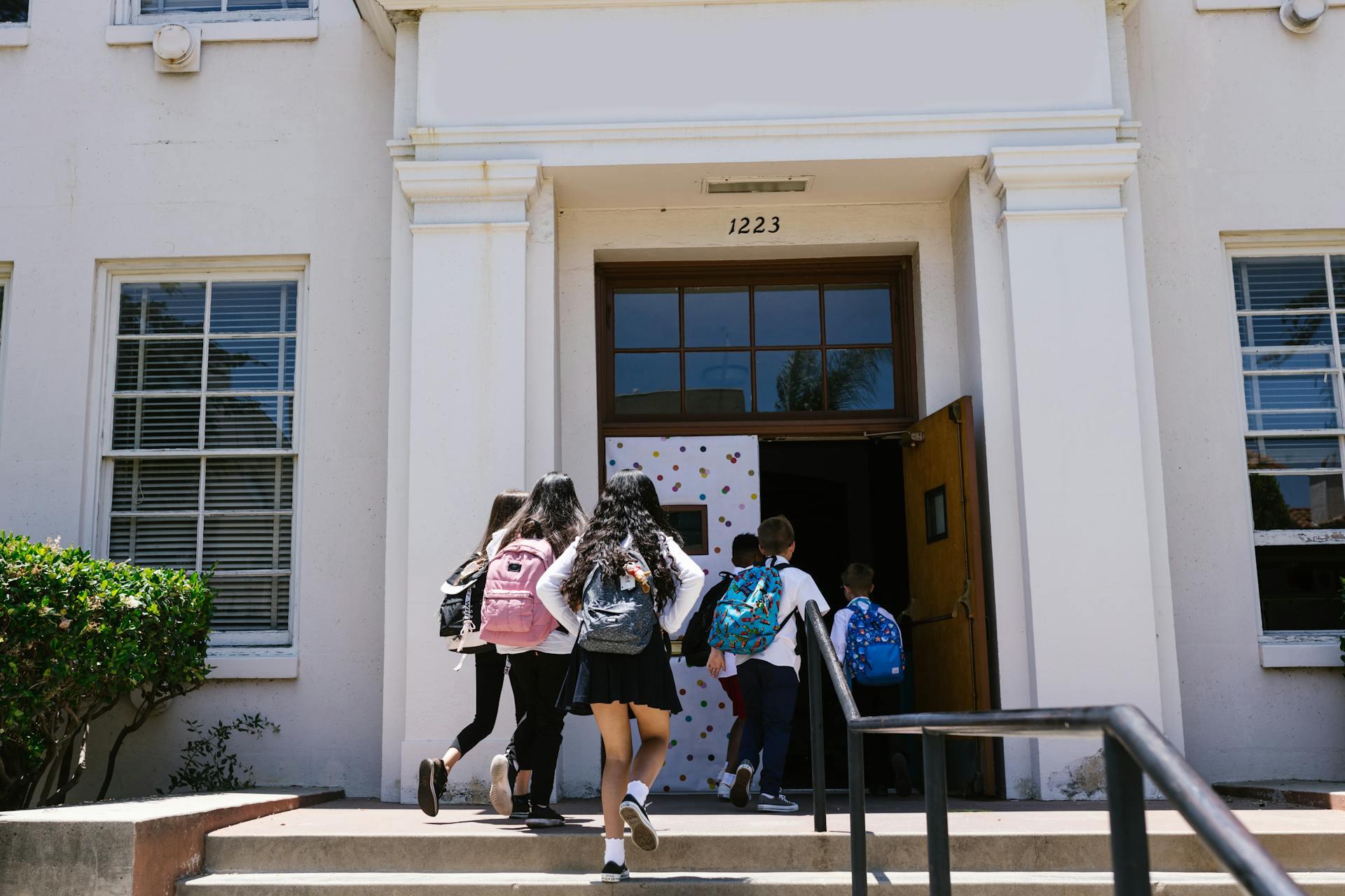 Group of school children with backpacks entering a building through open doors numbered 1223 on a sunny day.
