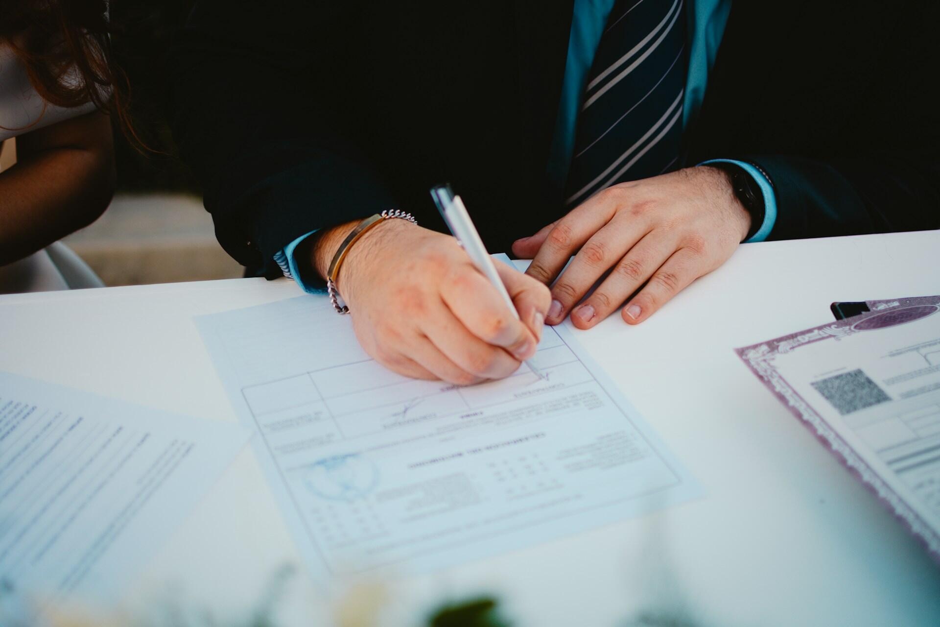 Person in formal attire signing a document on a white table with other papers nearby.