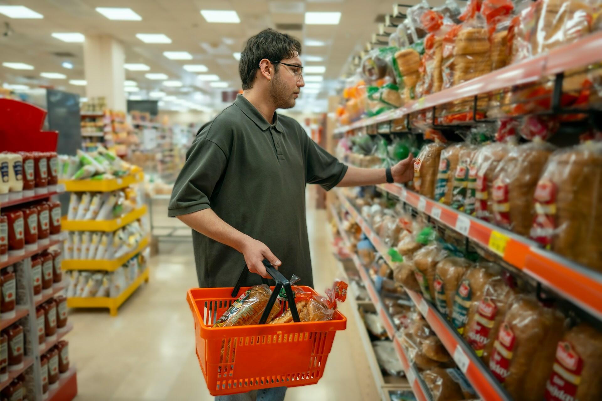A person carrying a red shopping basket, picking a sac of bread.