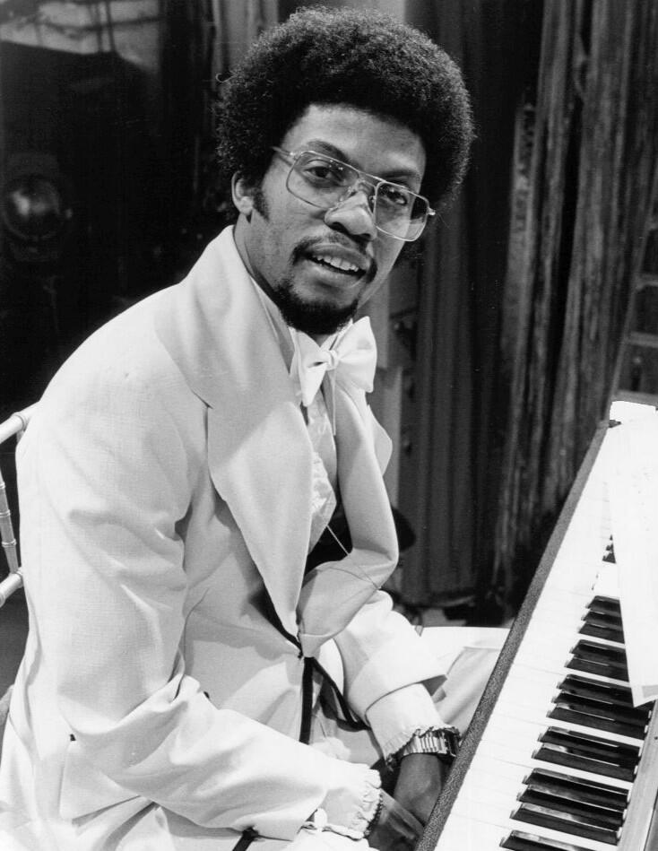 Black and white portrait of a smiling man with an afro and large glasses wearing a light suit and bow tie, seated next to a piano keyboard.
