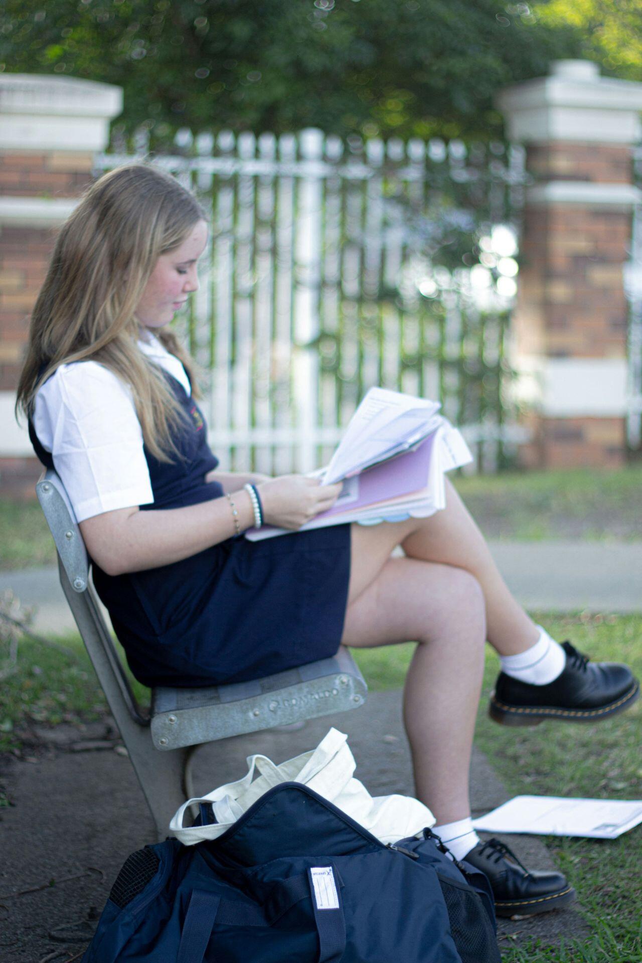 Student in uniform sitting on outdoor bench, reading papers with school bags on the ground beside her.