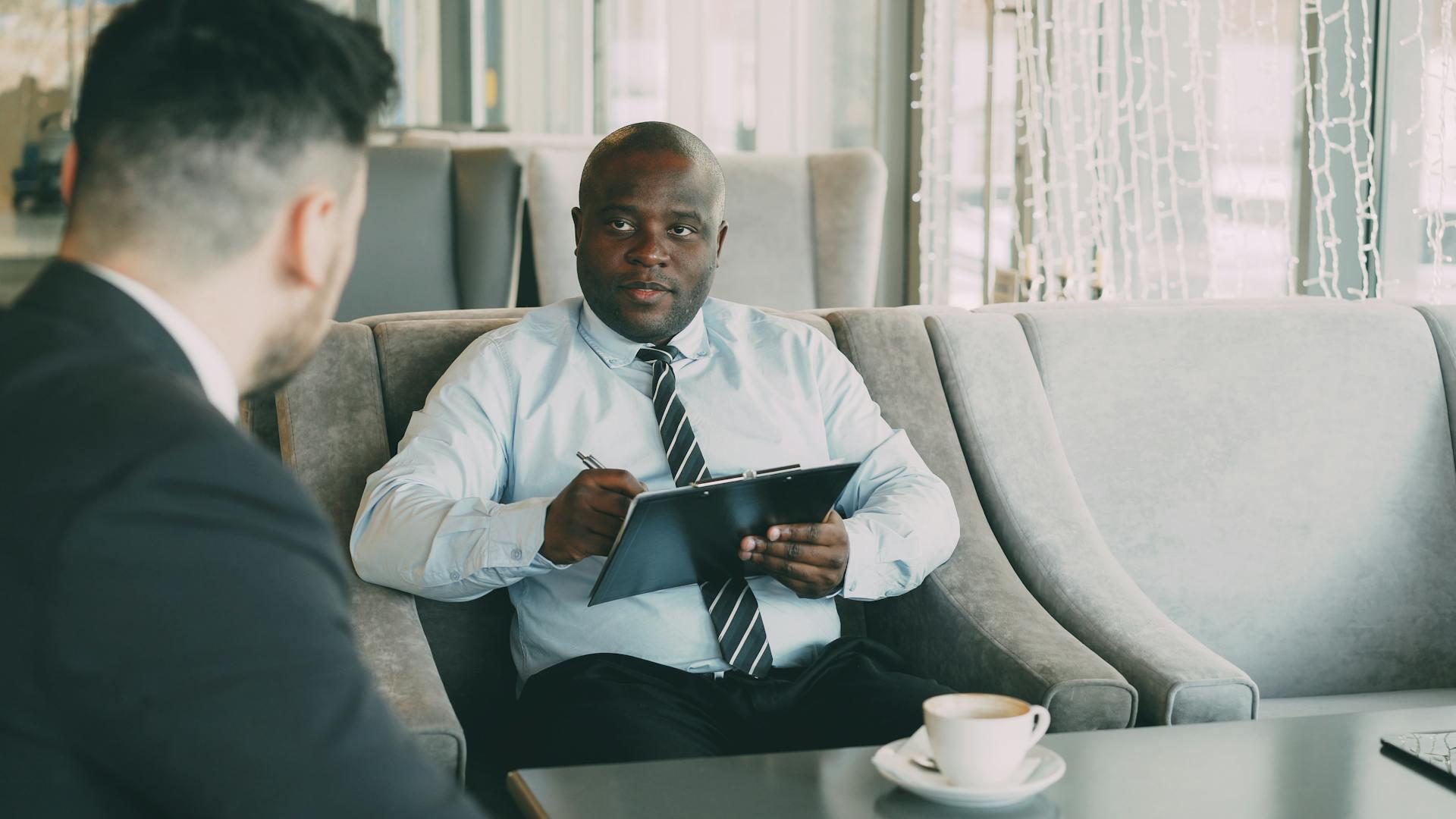 Two men in business attire sit in a lounge, one holding a clipboard and pen, the other facing him, with a coffee cup on the table.
