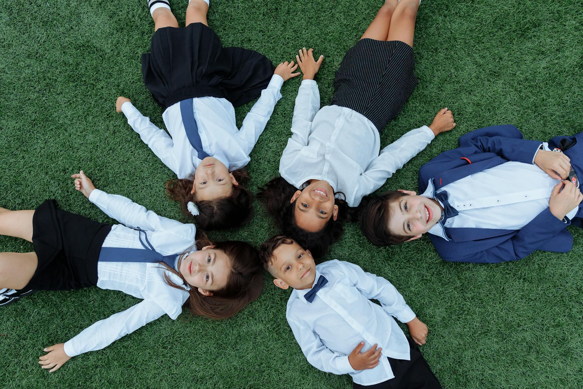Five children in school uniforms lying on grass in a circle with heads together and holding hands, seen from above.
