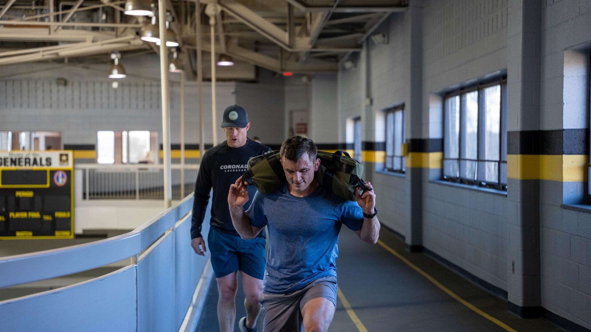 A person in a blue shirt carries a sandbag while doing lunges with another person following them.