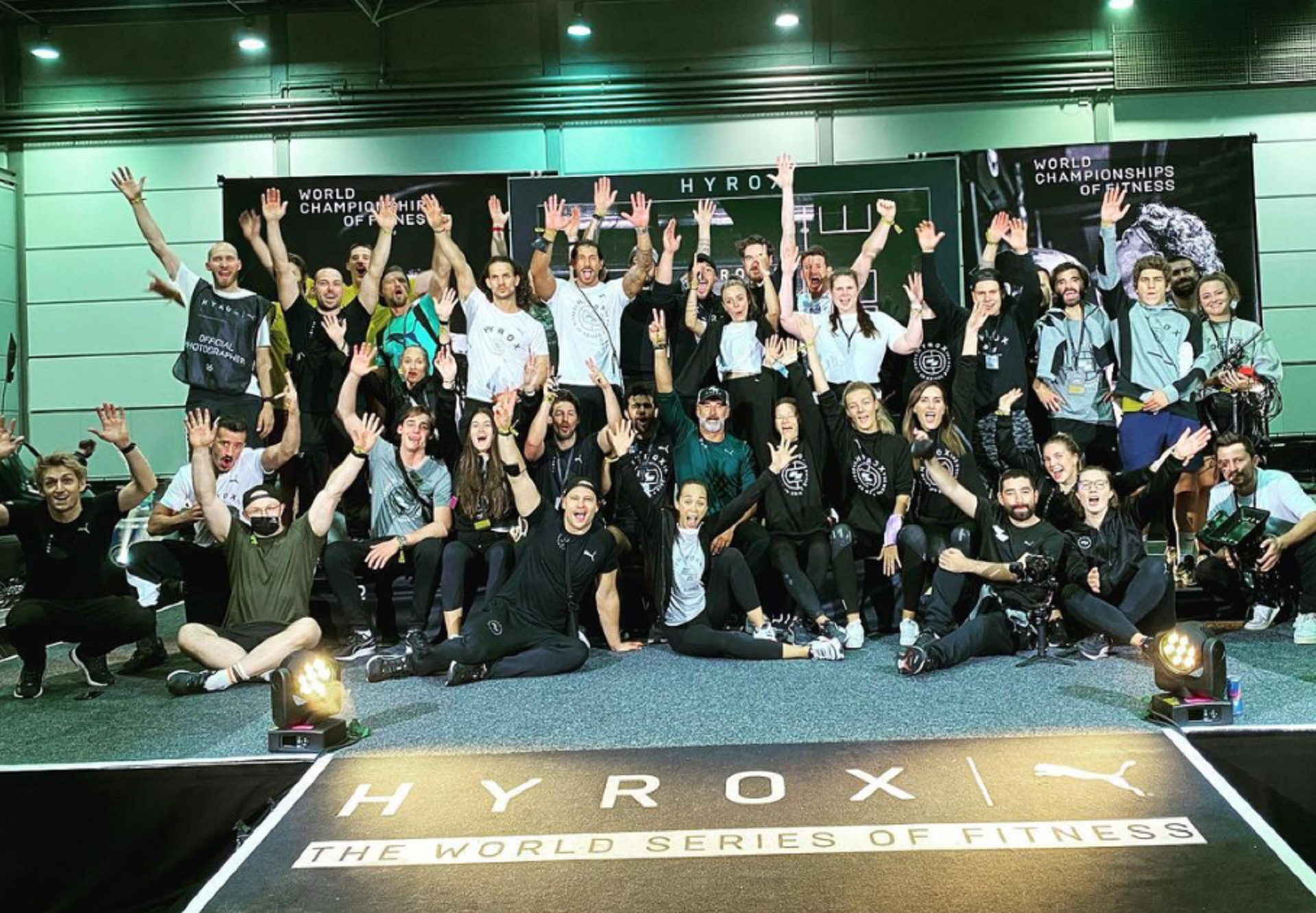 A large group of people posed with enthusiastic expresstions in front of a black and white sign.