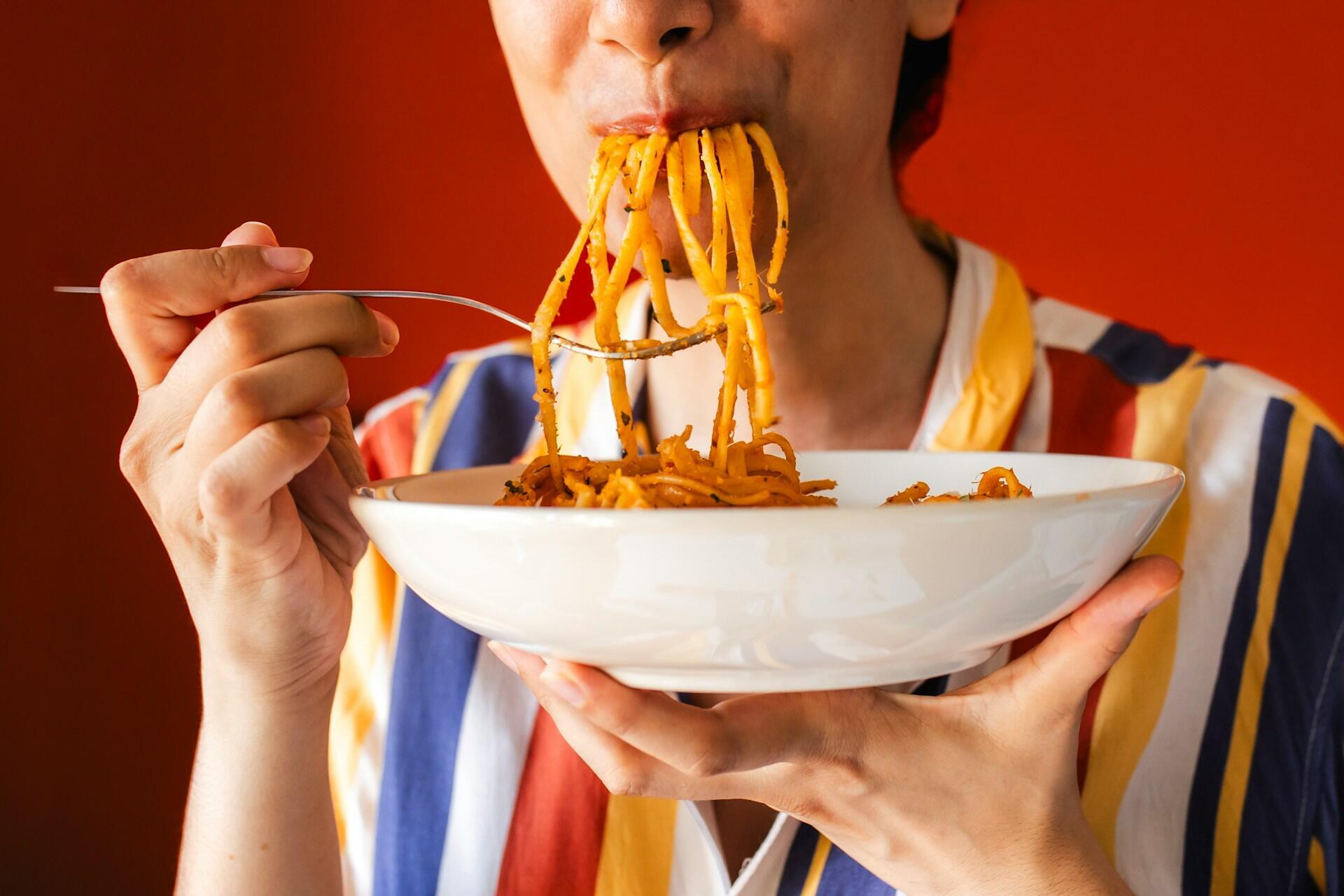 A person in a striped shirt, holding a food bowl, with noodles hanging out of their mouth.