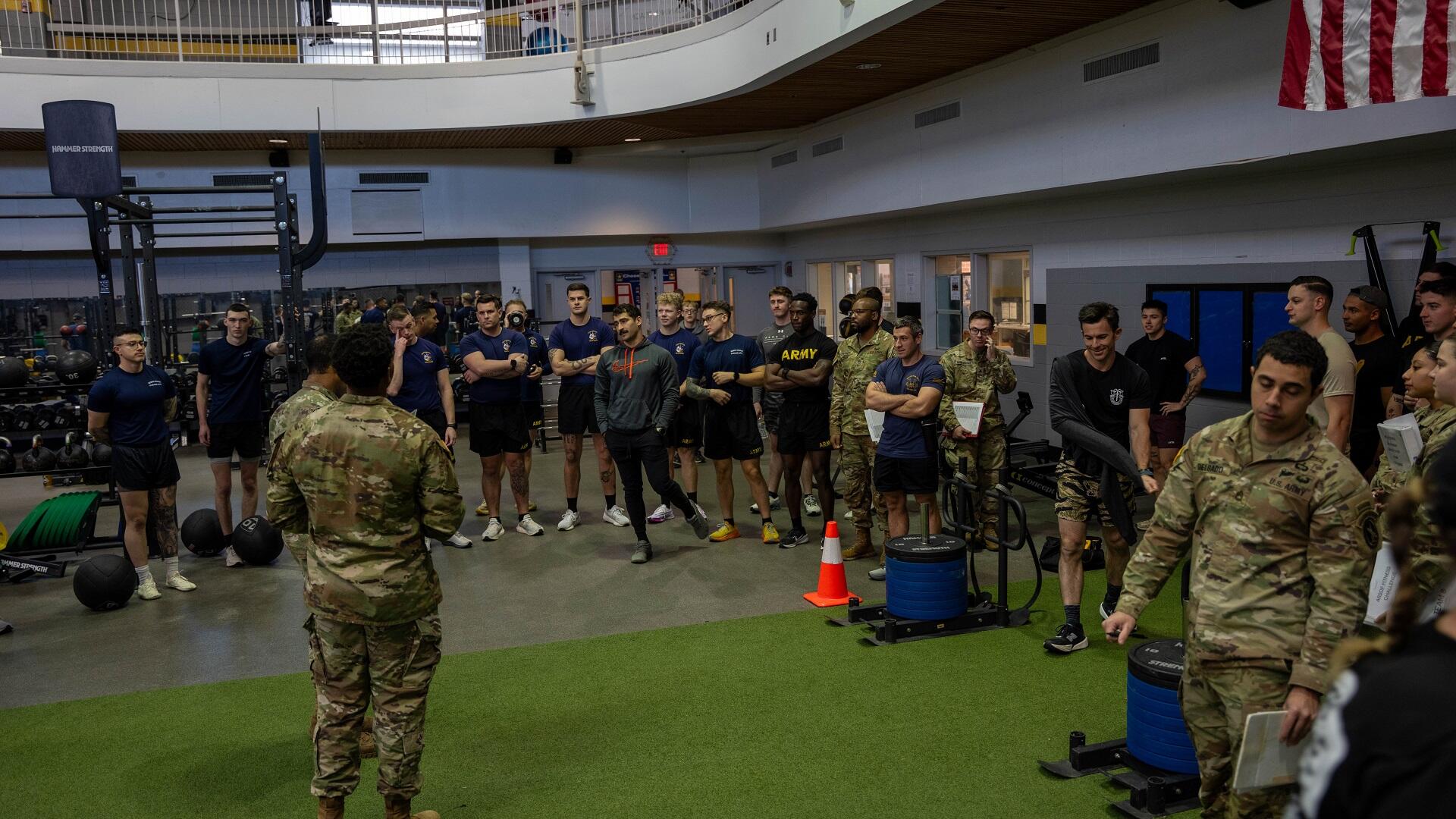 A group of people, some in athletic wear and others in military uniform, stand around in a fitness training facility.