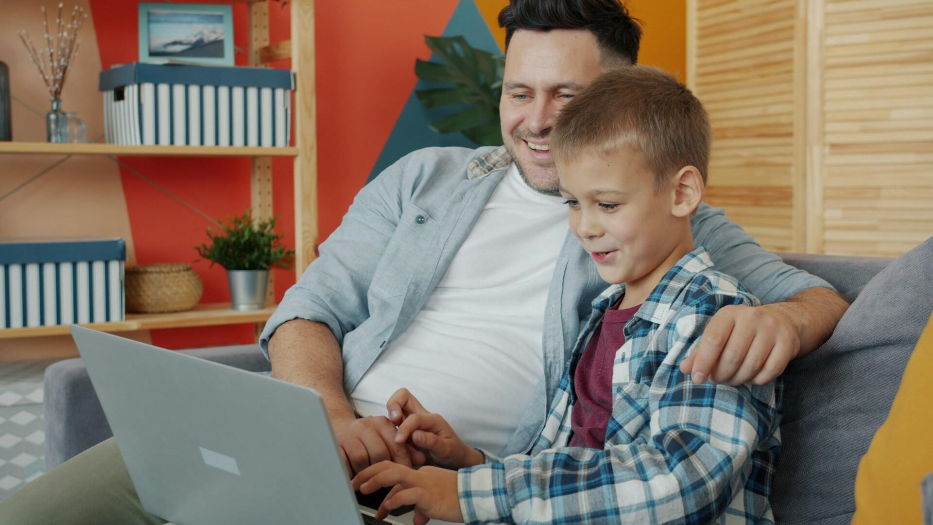 Man and boy sitting on a couch using a laptop together in a cozy, colorful living room.
