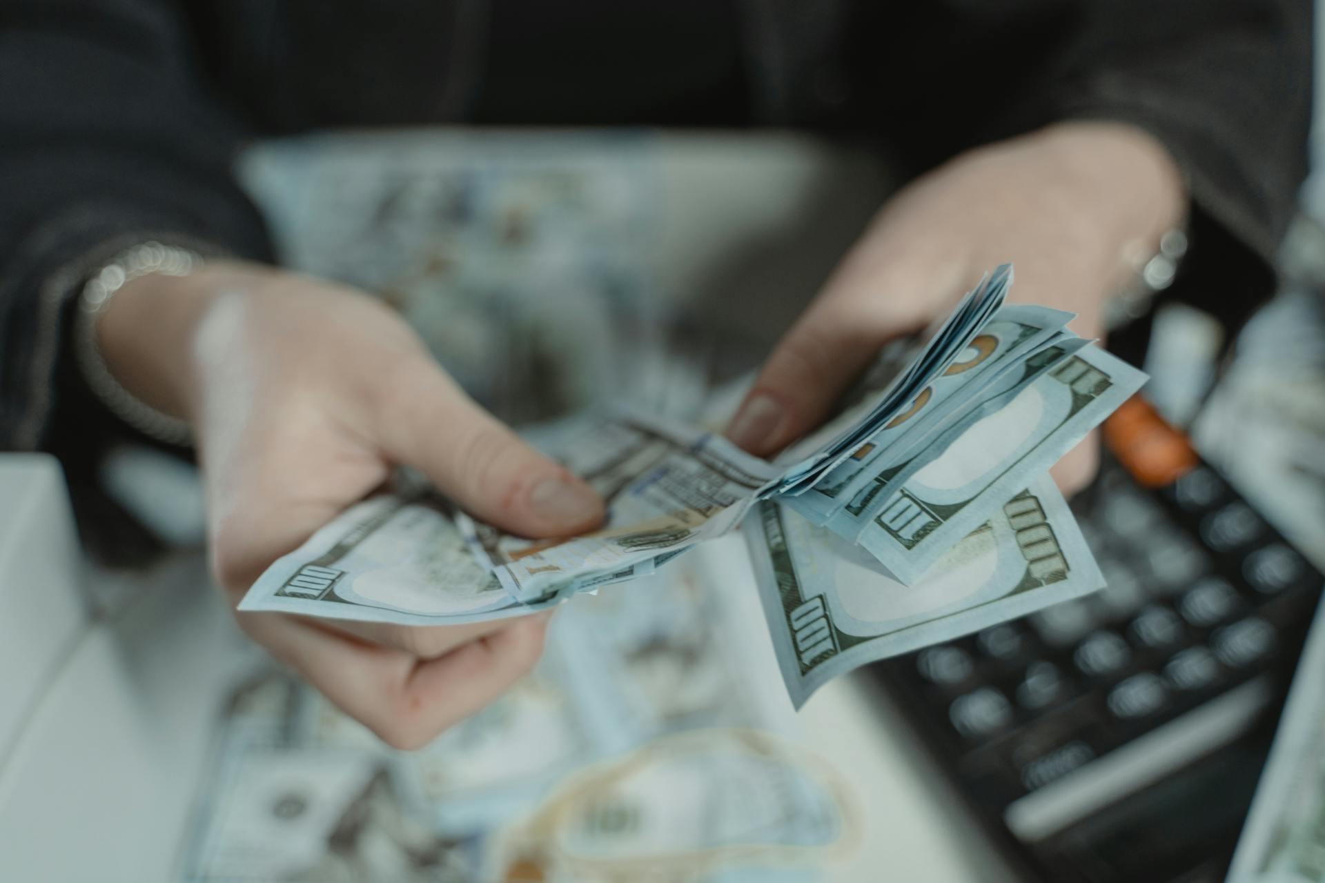Person counting multiple US hundred-dollar bills with a calculator nearby on the table.