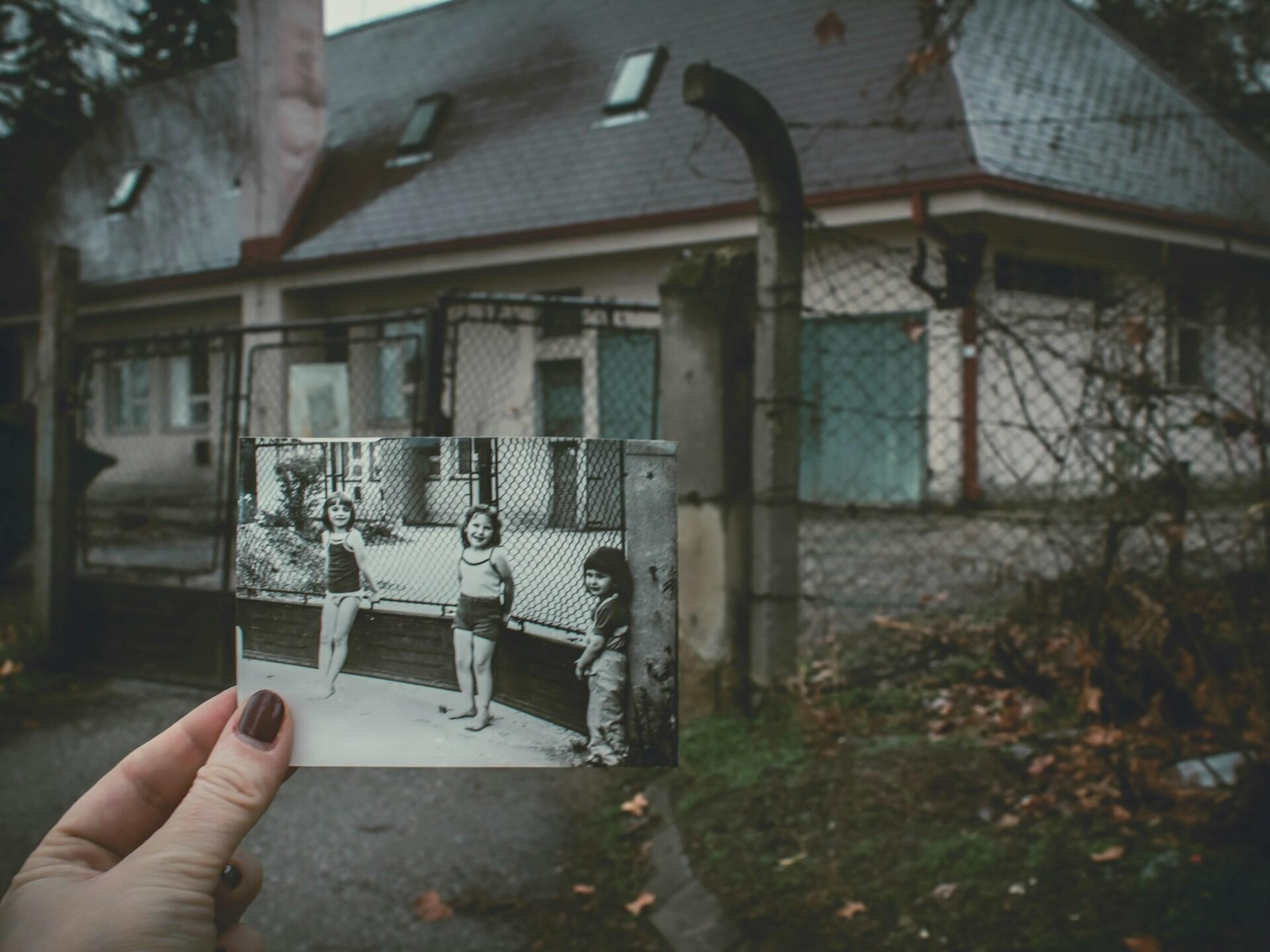 A person holds up a picture of three small girls in front of an old house.