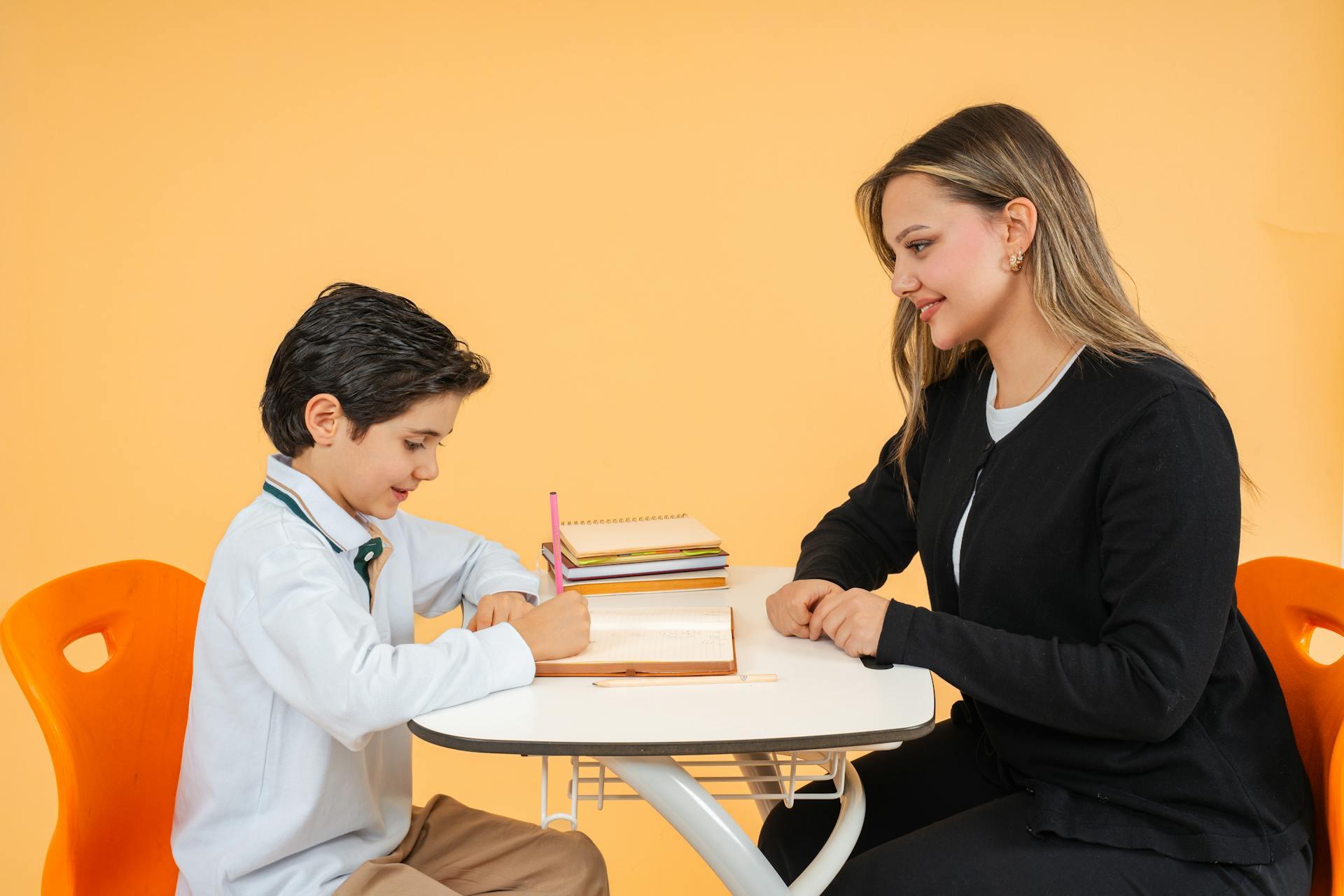 Teacher assisting a student with writing at a desk in a bright, orange-themed classroom setting.