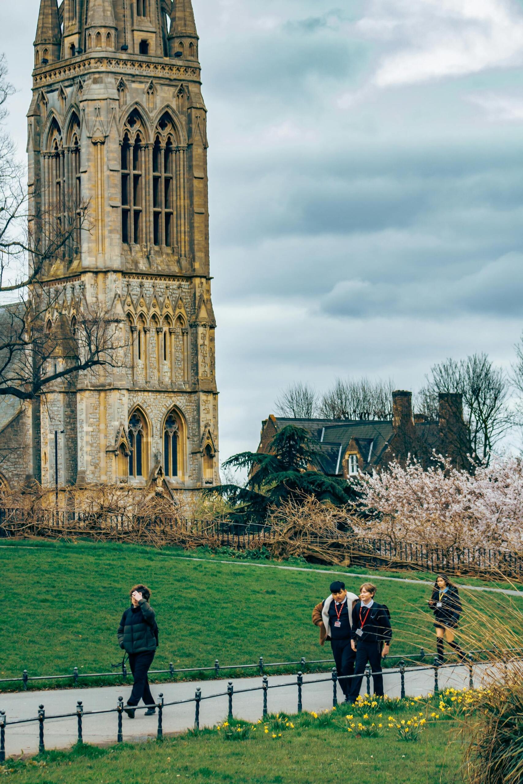 People walking on a path near an old Gothic stone tower with blossoming trees and houses in the background under a cloudy sky.