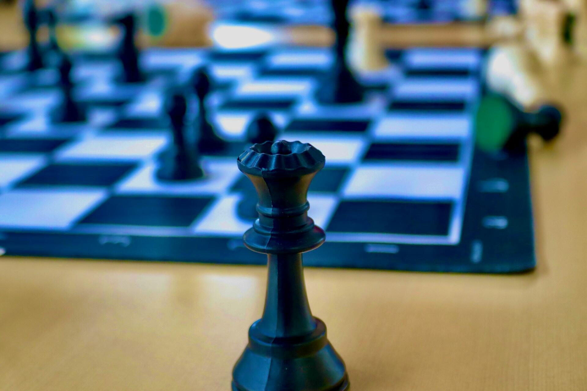 Close-up of a black queen chess piece on a chessboard with other pieces blurred in the background