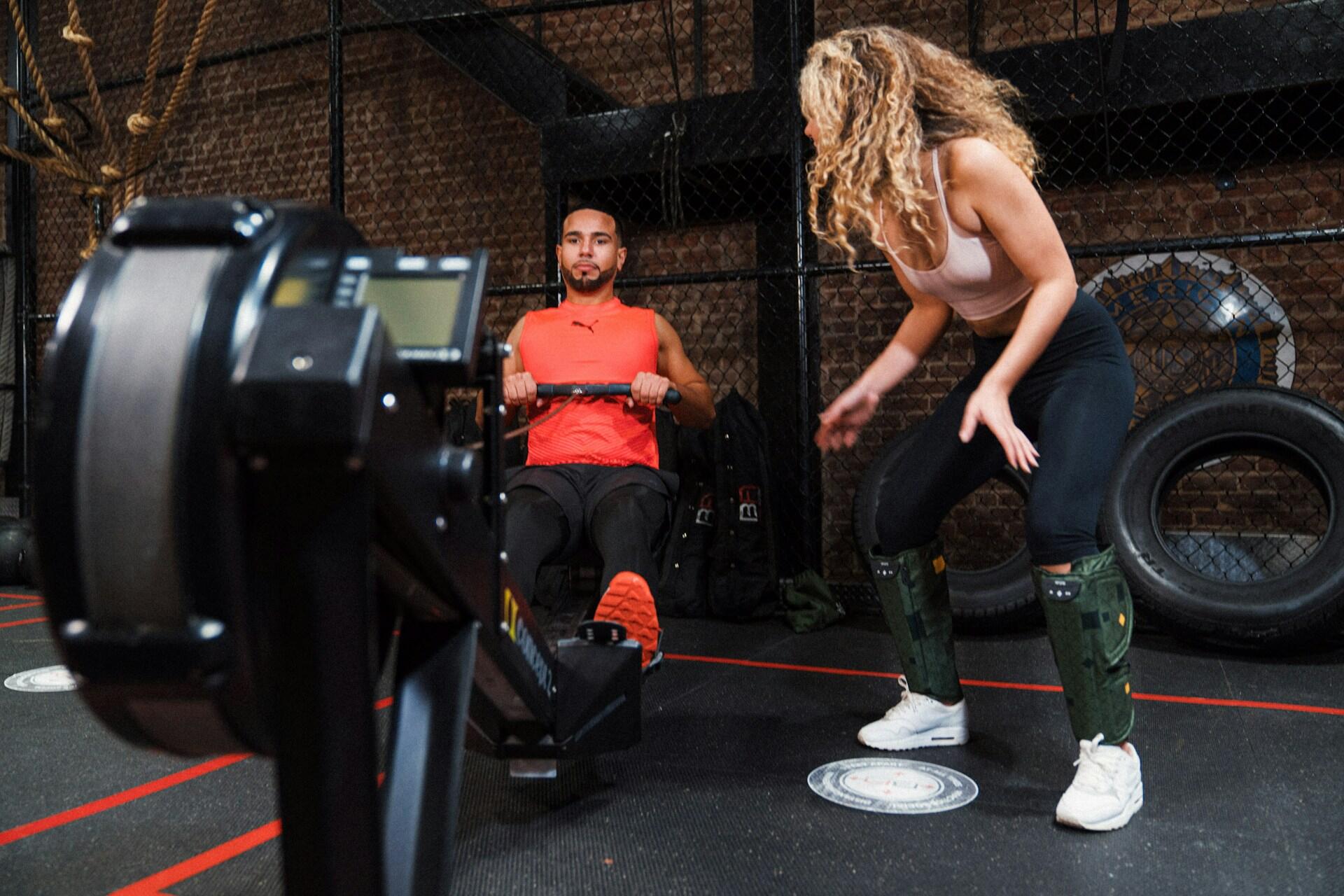 A man working a rowing machine in a training facility while a woman wearing athletic gear looks on.