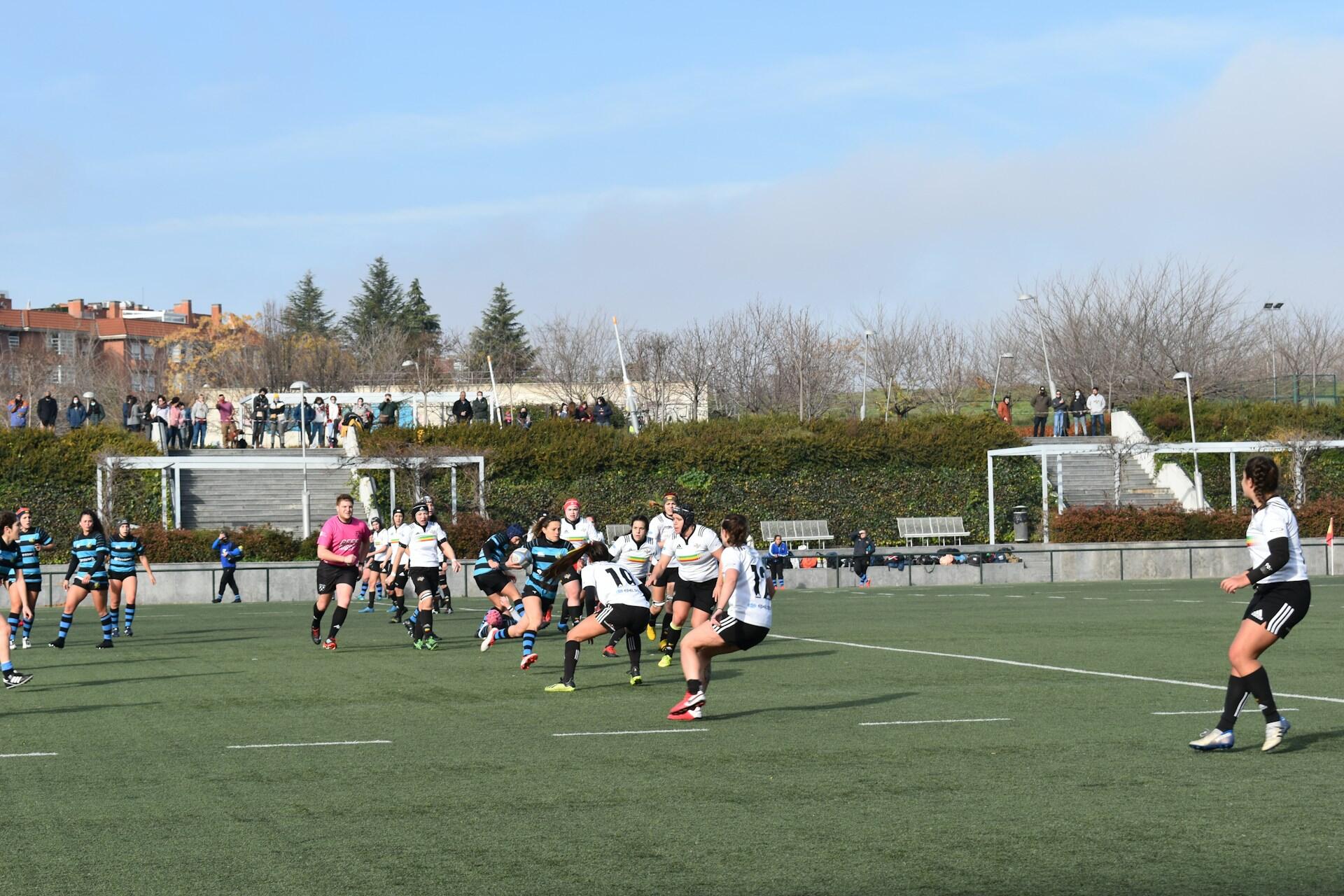 Women’s rugby match in progress on a green field with players from two teams and spectators in the background.