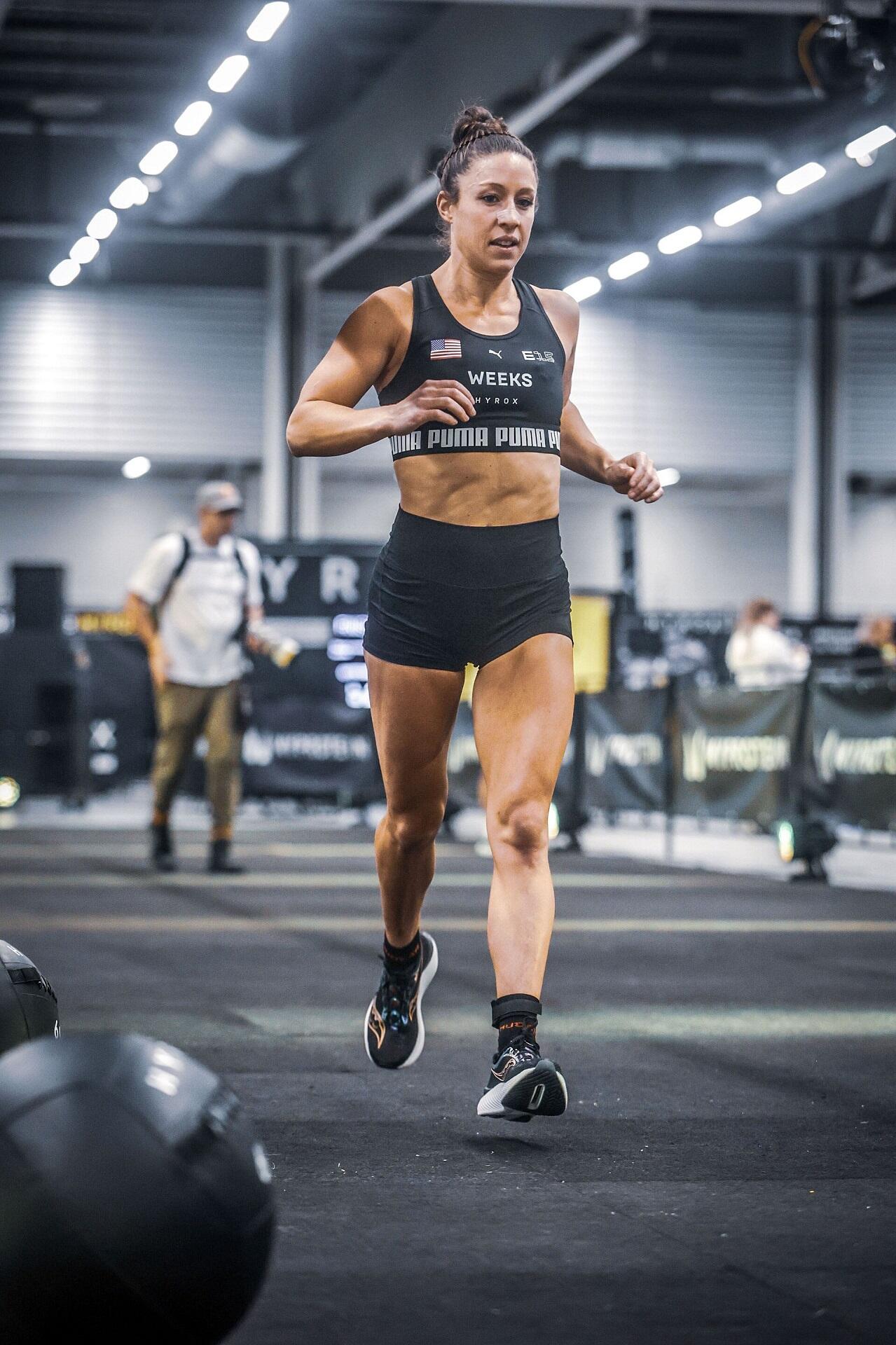 A person wearing black athletic clothing runs on an indoor track surrounded by gym equipment.