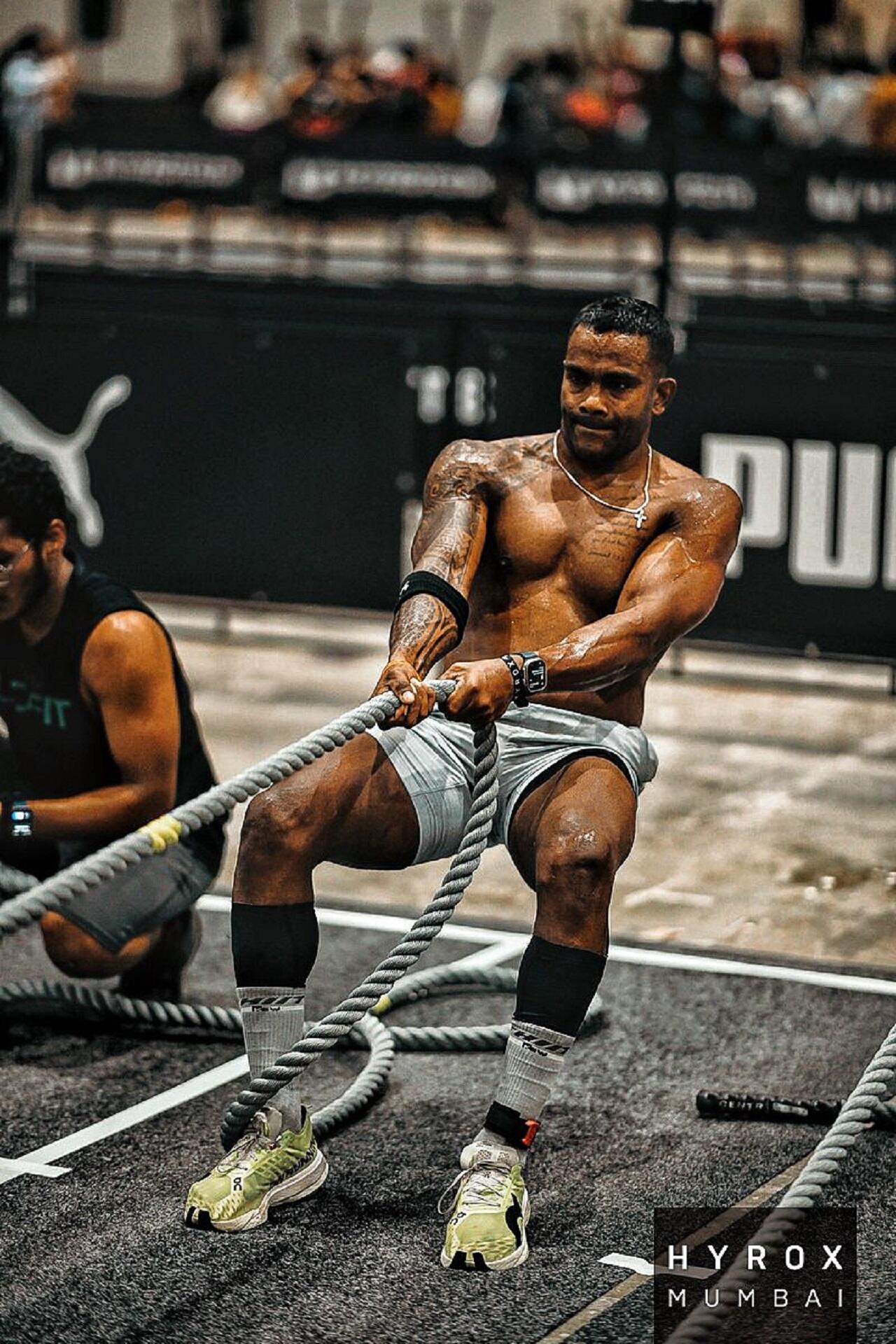 A bare-chested athlete pulls hard on a rope in an indoors space.