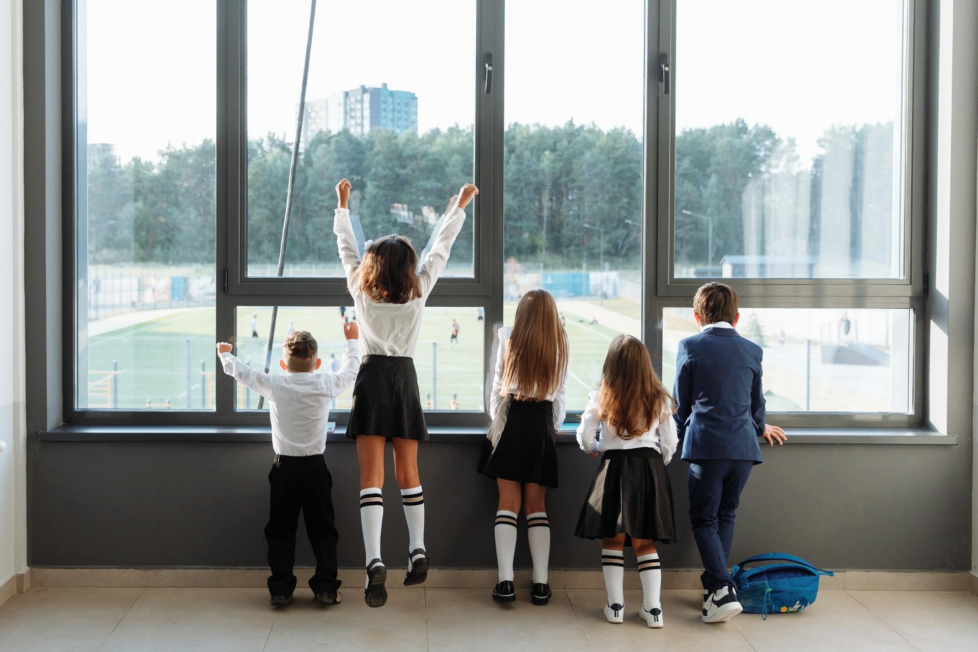 Five schoolchildren in uniform stand with their backs to the camera looking out large bright windows overlooking a green outdoor area.