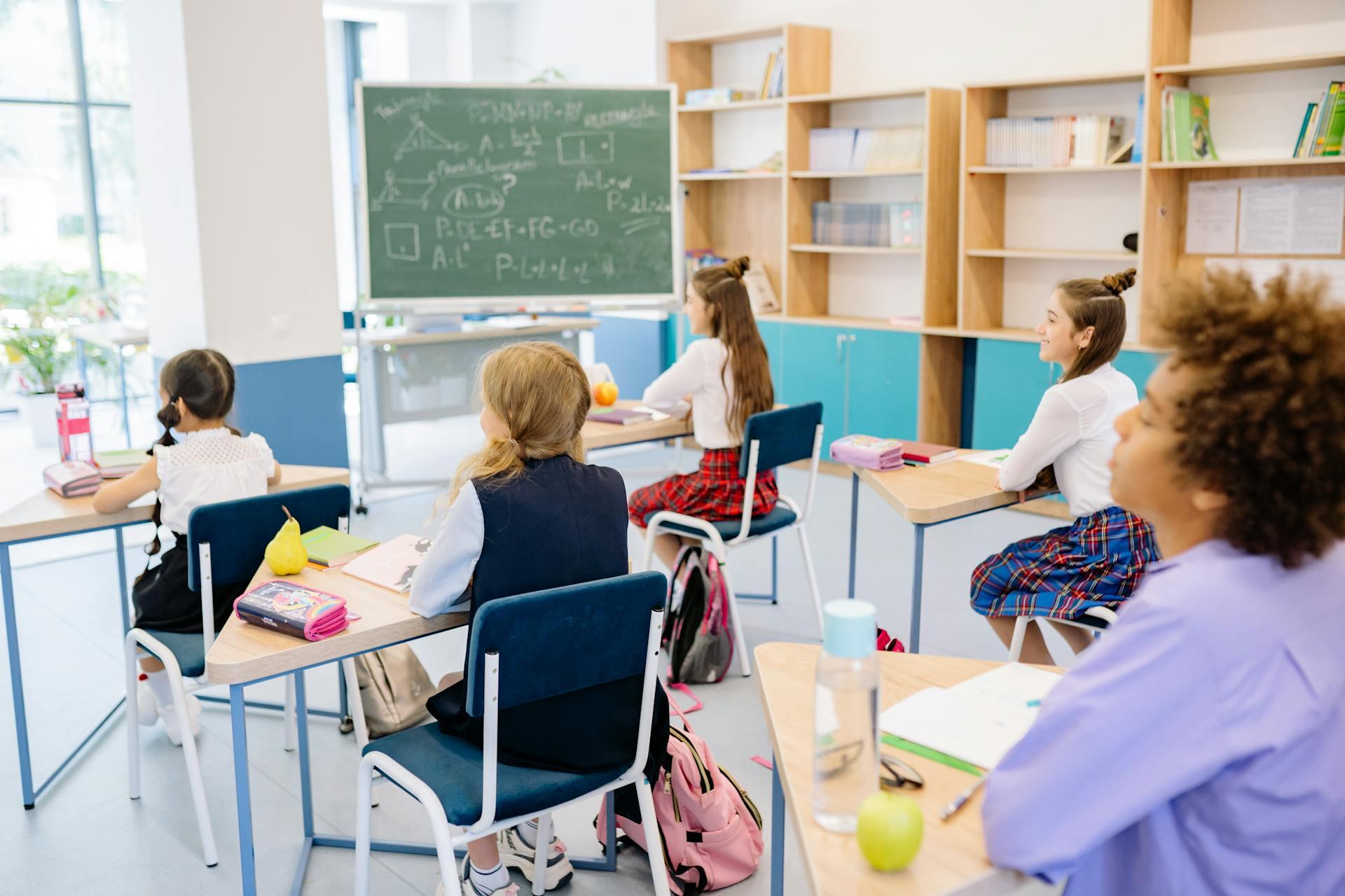 Students seated at desks in a classroom focused on a chalkboard with math equations, surrounded by bookshelves and school supplies.