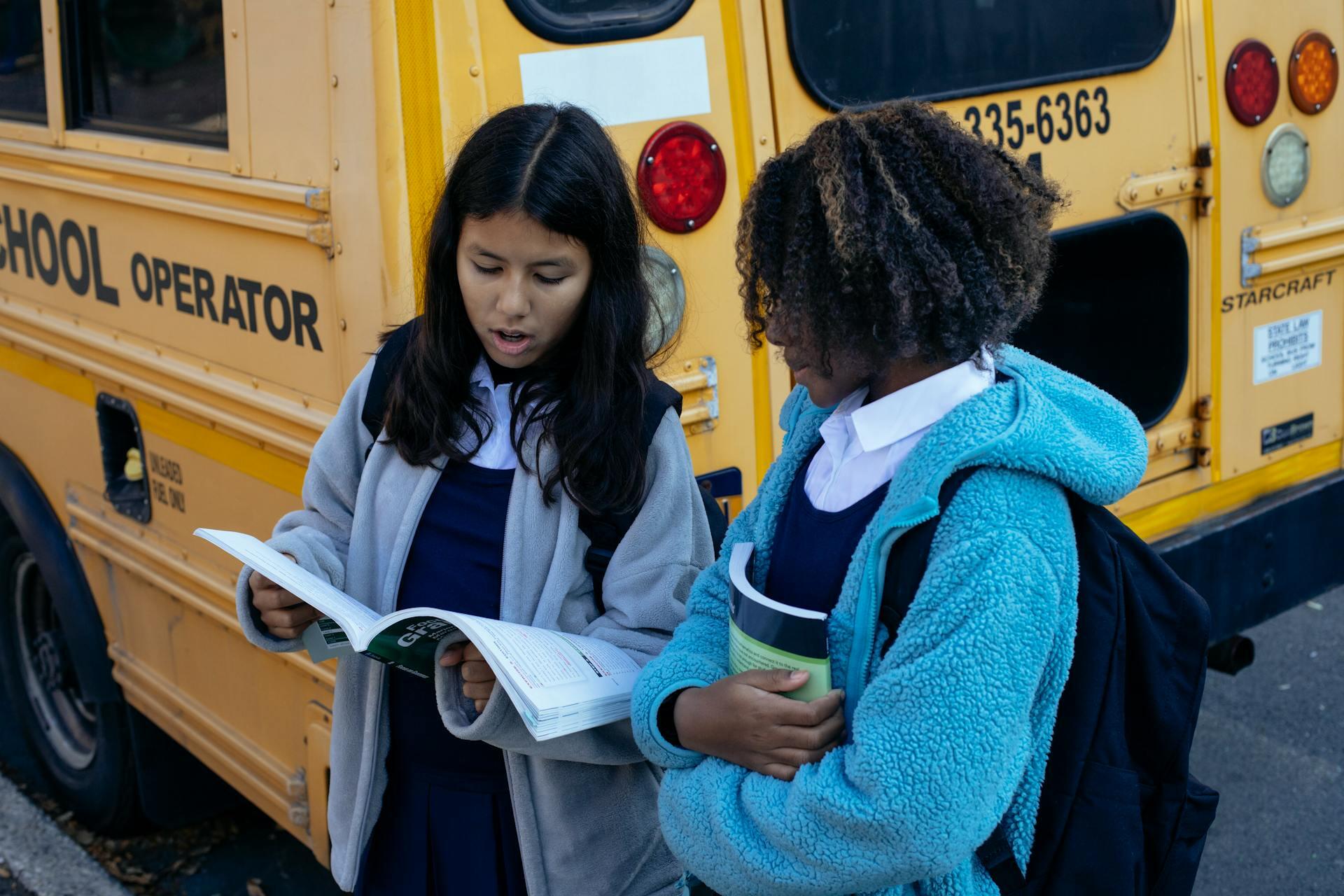 Two schoolgirls in uniforms stand by a yellow school bus, one reading a book and the other holding books, both wearing jackets and backpacks.