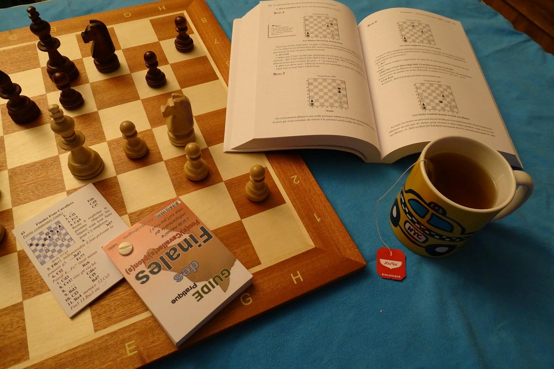 A chessboard set beside an open chess book, analysis cards, and a cup of tea while studying chess tactics.