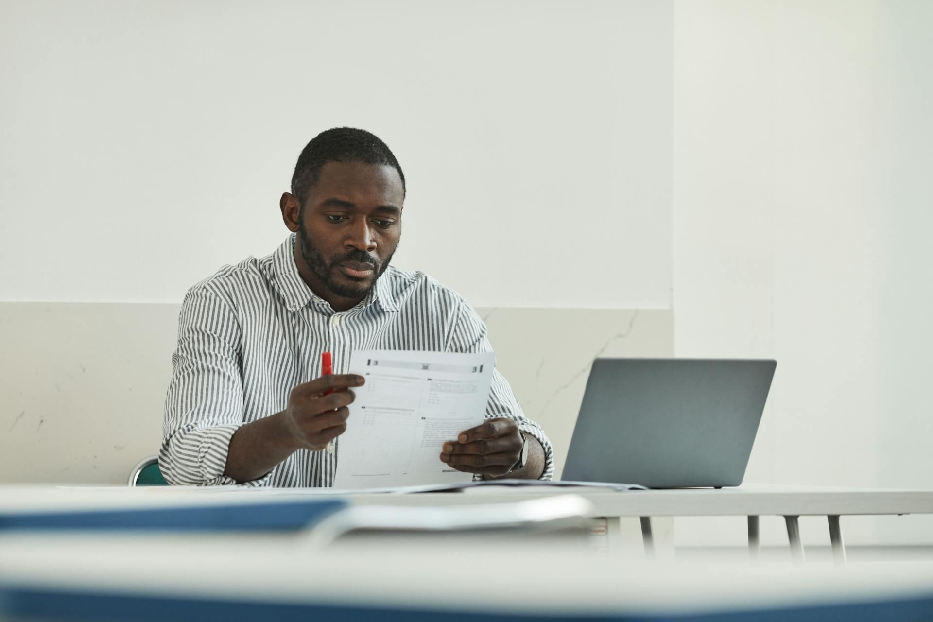 Man in a striped shirt reviewing a document with a red pen, seated at a table with a closed laptop in a minimalistic room.
