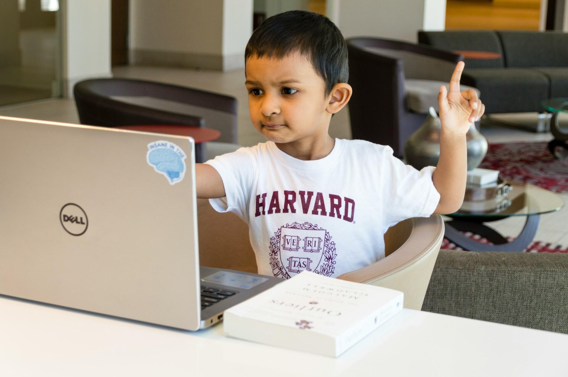 A child wearing a white tee-shirt sits in front of a computer with their hand raised.