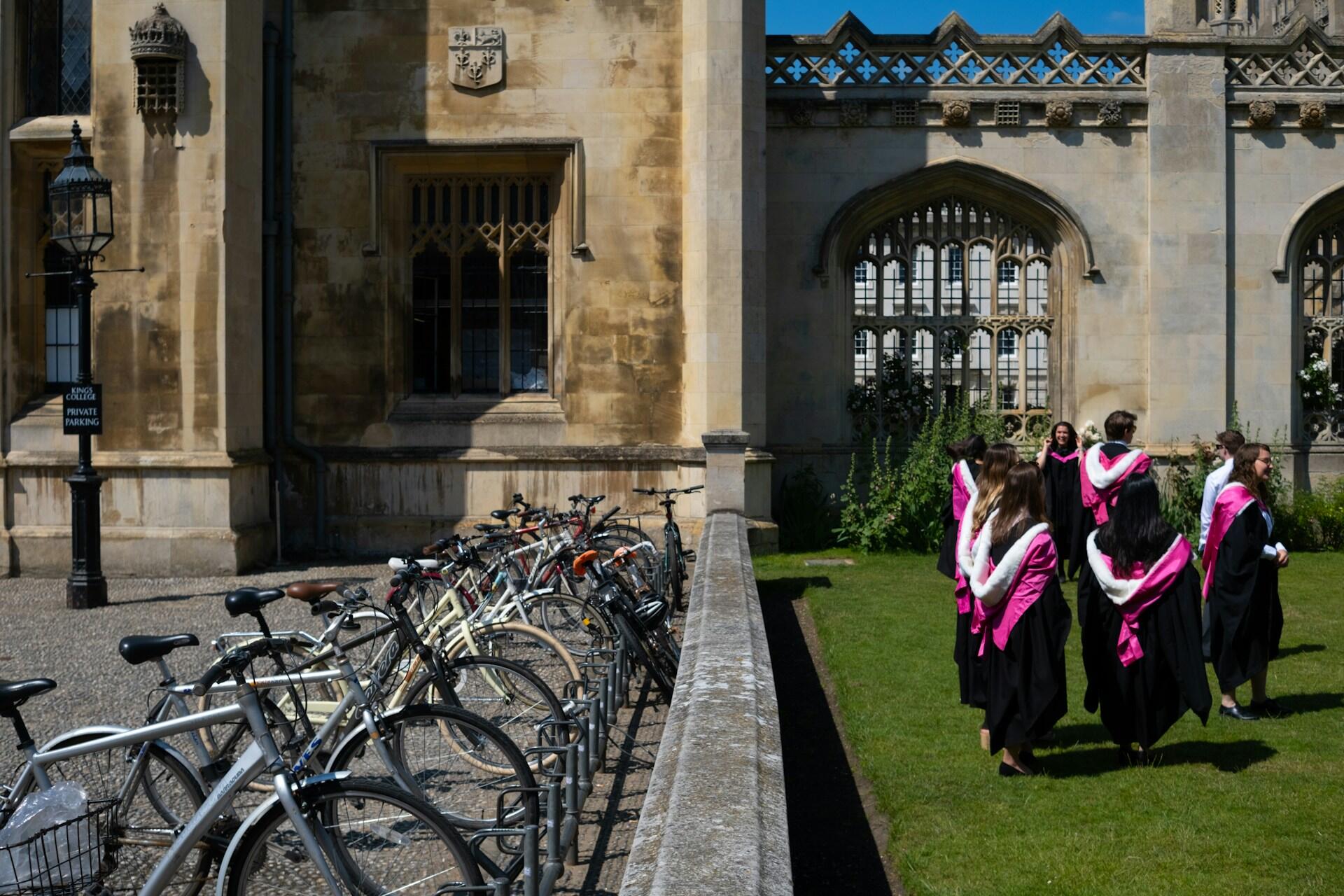 Group of graduates in black gowns with pink hoods standing on a grassy courtyard beside a row of parked bicycles by a historic stone building.