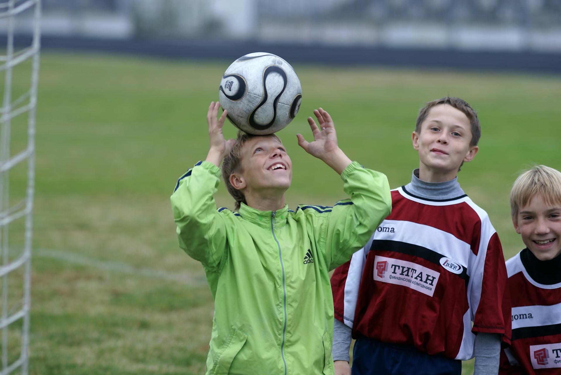 Children playing soccer near goalpost, one boy in green jacket heading the ball, others in red and white jerseys watching.