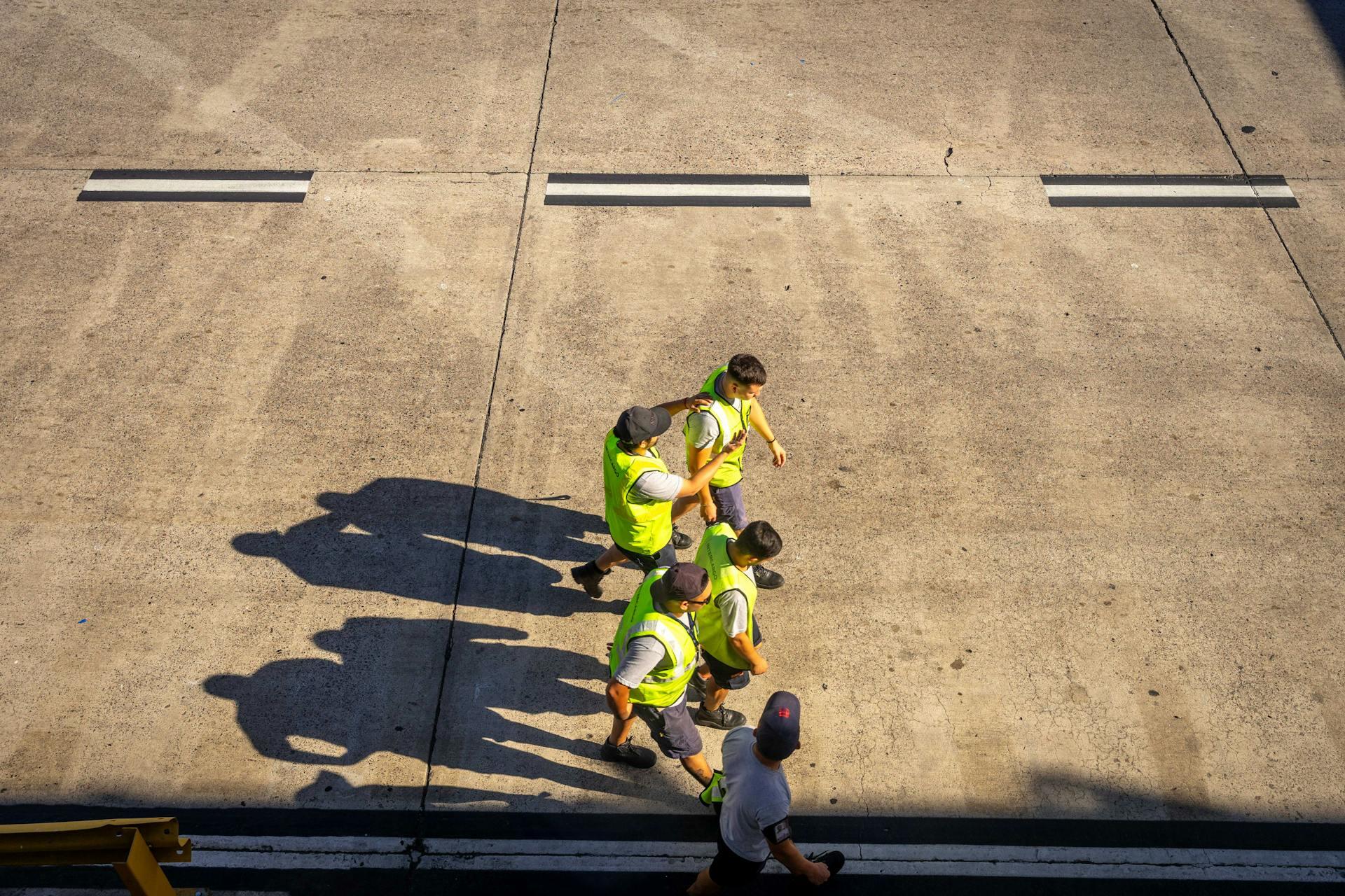 Aerial view of four workers in yellow vests and one person walking on a concrete surface with marked lines and shadows.