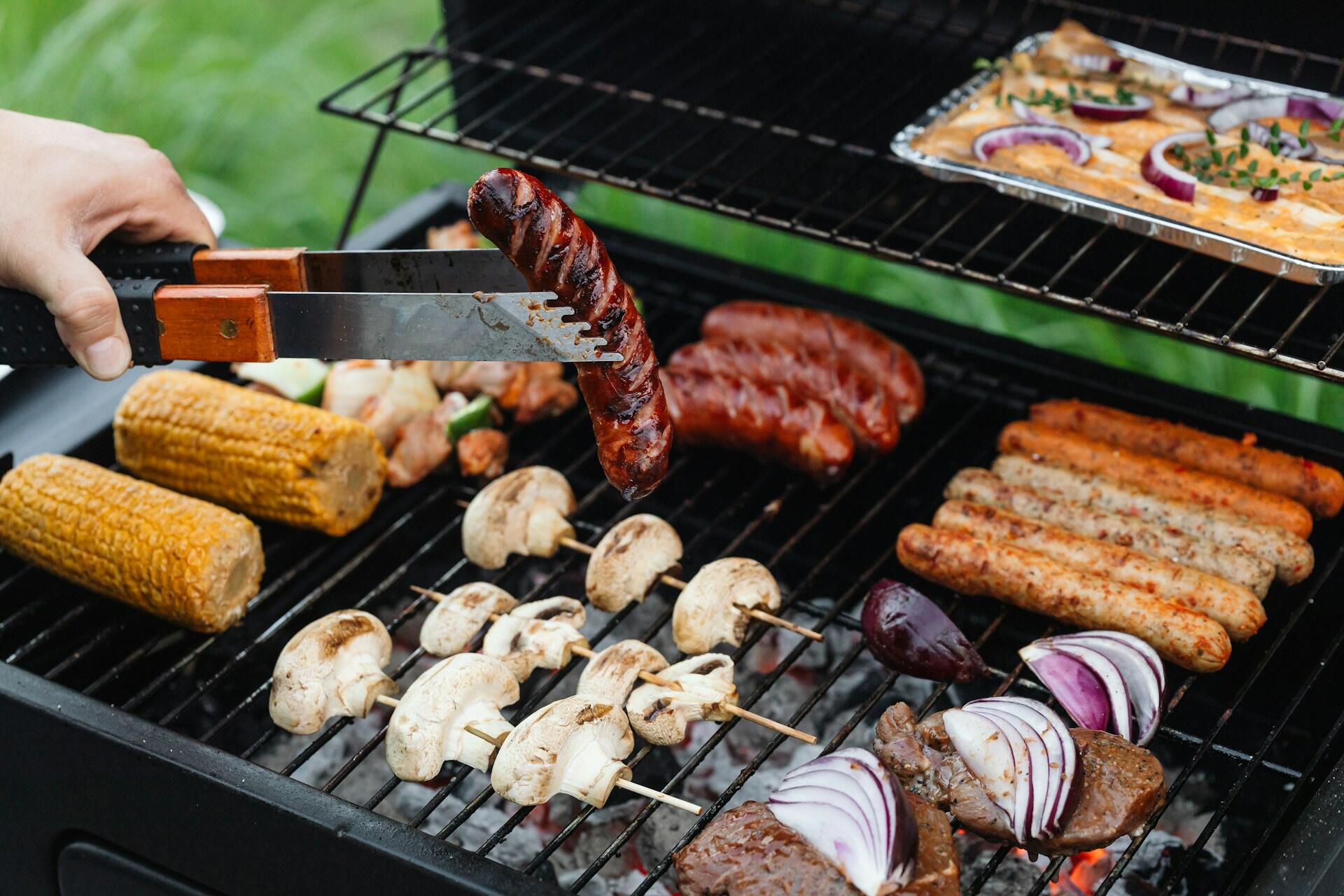 A barbecue grill loaded with grilled meats and veg with someone holding a grilled sausage with a pair of tongs over it.