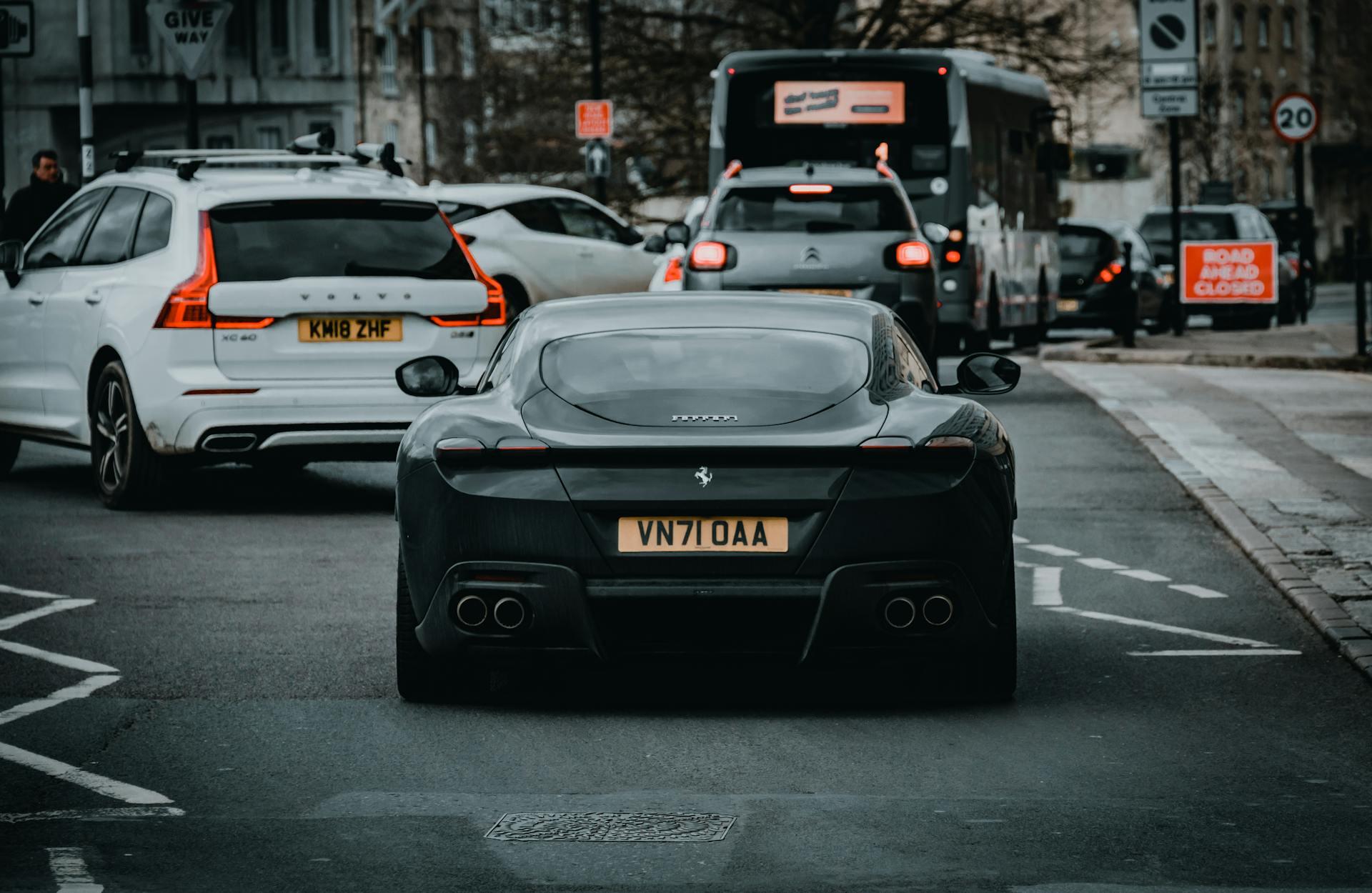 Rear view of a black Ferrari sports car on a city street surrounded by other vehicles including a white Volvo and a bus.