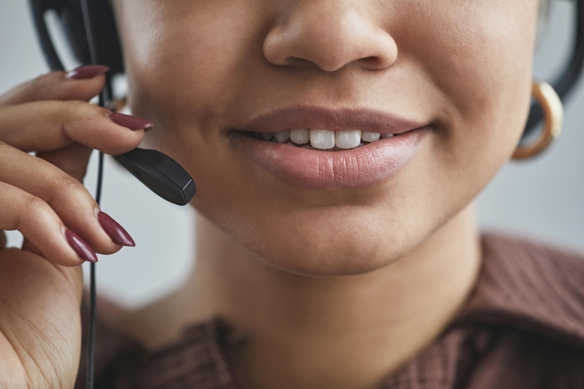 Close-up of a smiling woman wearing a headset, adjusting the microphone boom with a manicured hand.