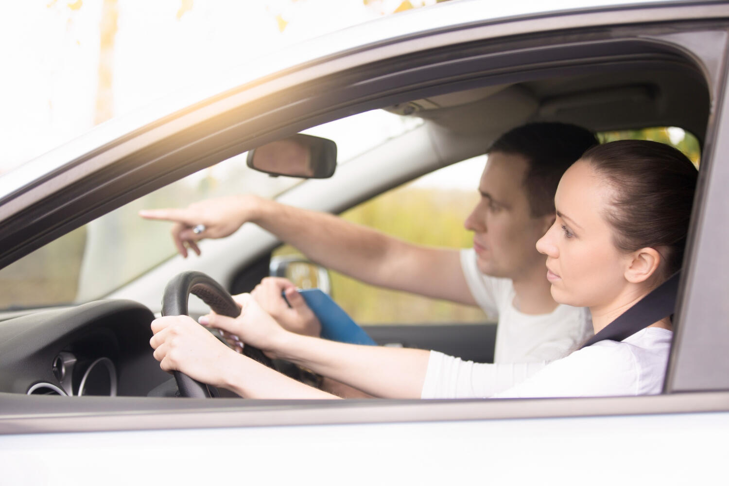 Driving instructor pointing ahead while student drives a car, both wearing seat belts.