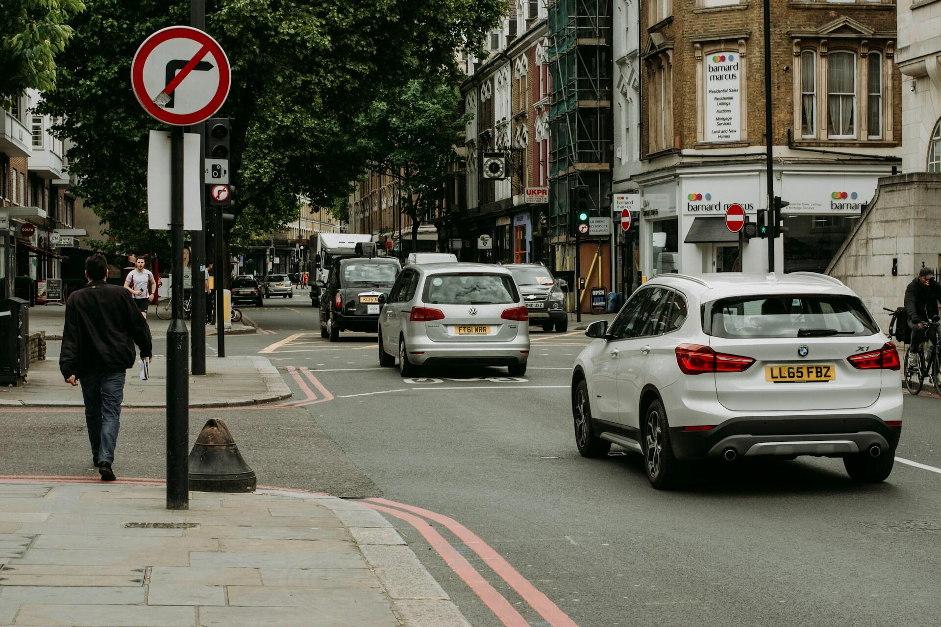 Urban street scene with cars driving, pedestrians walking, and a no right turn sign visible on a pole.