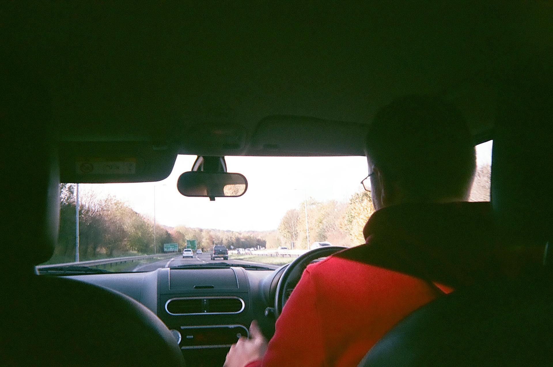 View from the backseat showing a person in a red hoodie driving a car on a highway with trees along the road.